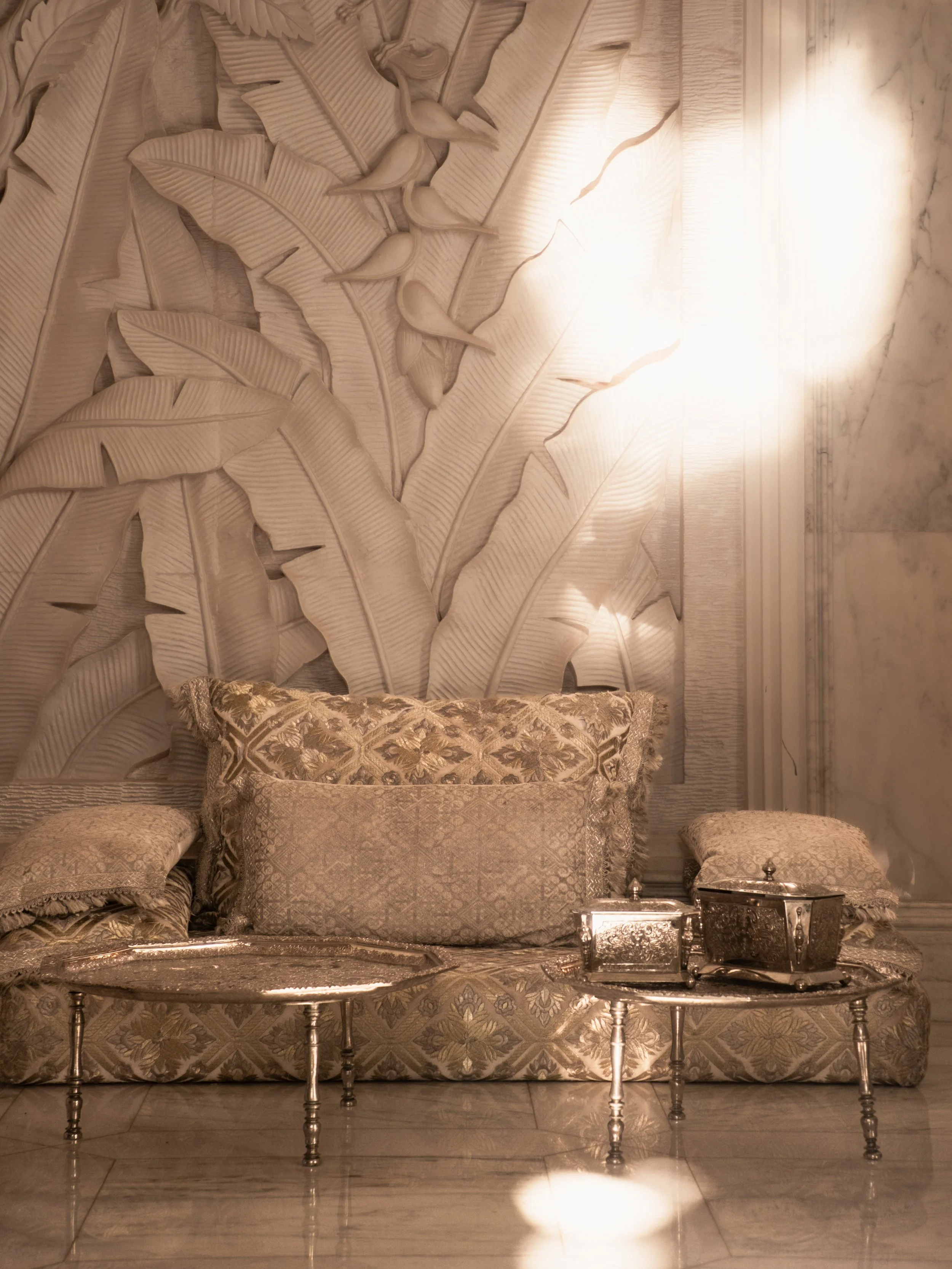 An ornate silver table with a large, decorative silver tray, set in front of a plush sofa with patterned cushions, against a wall with intricate banana leaf carvings and white marble accents.