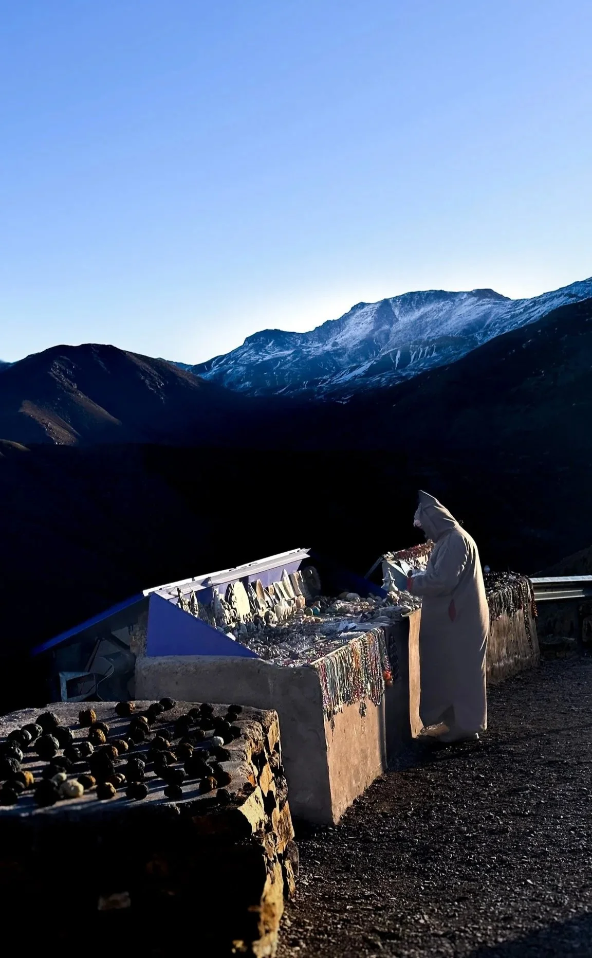 Person in a white coat shopping for jewelry at a roadside stall with mountains in the background.