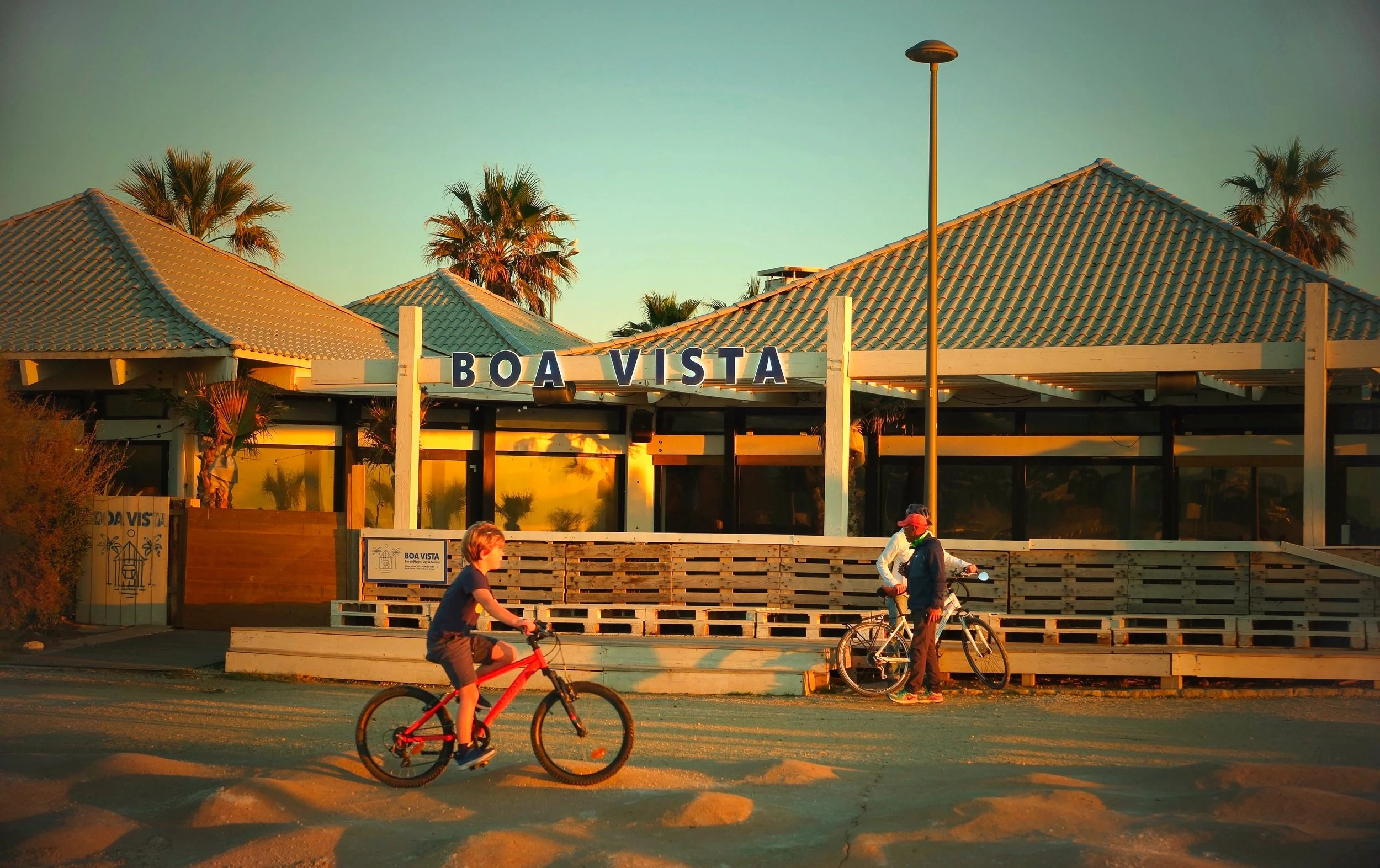 People riding bicycles in front of a building with a sign that says 'Boa Vista', palm trees, and a streetlight, during sunset.