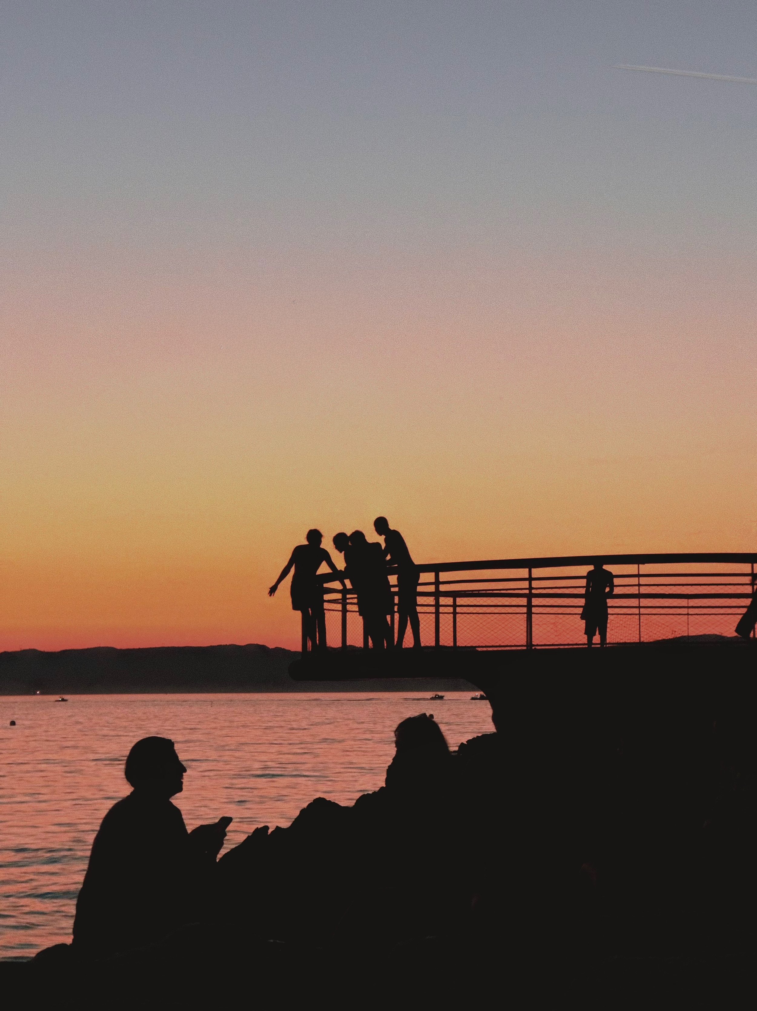 Silhouettes of people on a pier and rocks against a colorful sunset over a body of water.