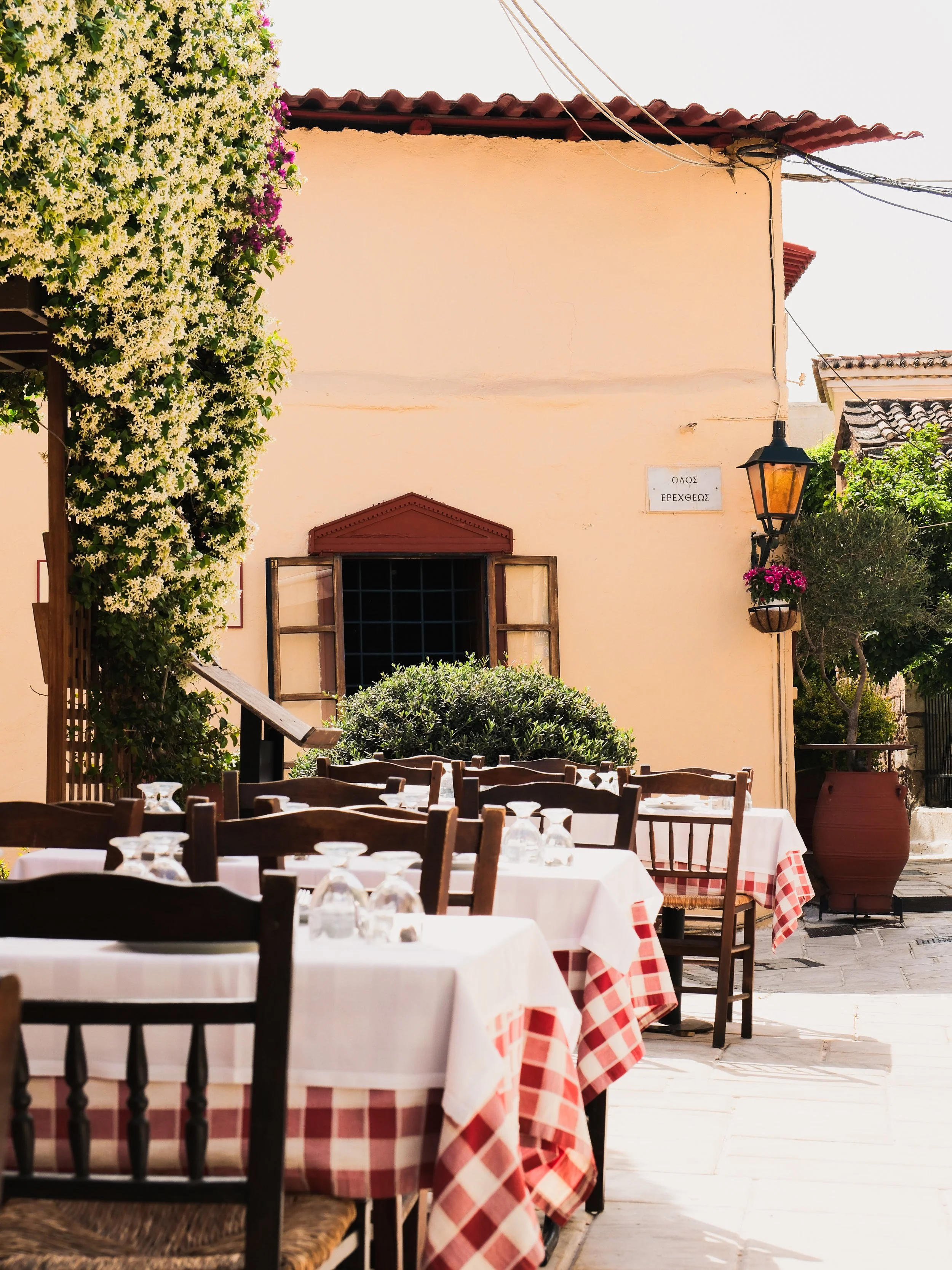 Outdoor restaurant with white tablecloths and red-checkered tablecloths, wooden chairs, and a building with pink walls, a red tiled roof, and blooming flowers, including a large vine with white and purple flowers, a street lamp, and a street sign with Greek writing.