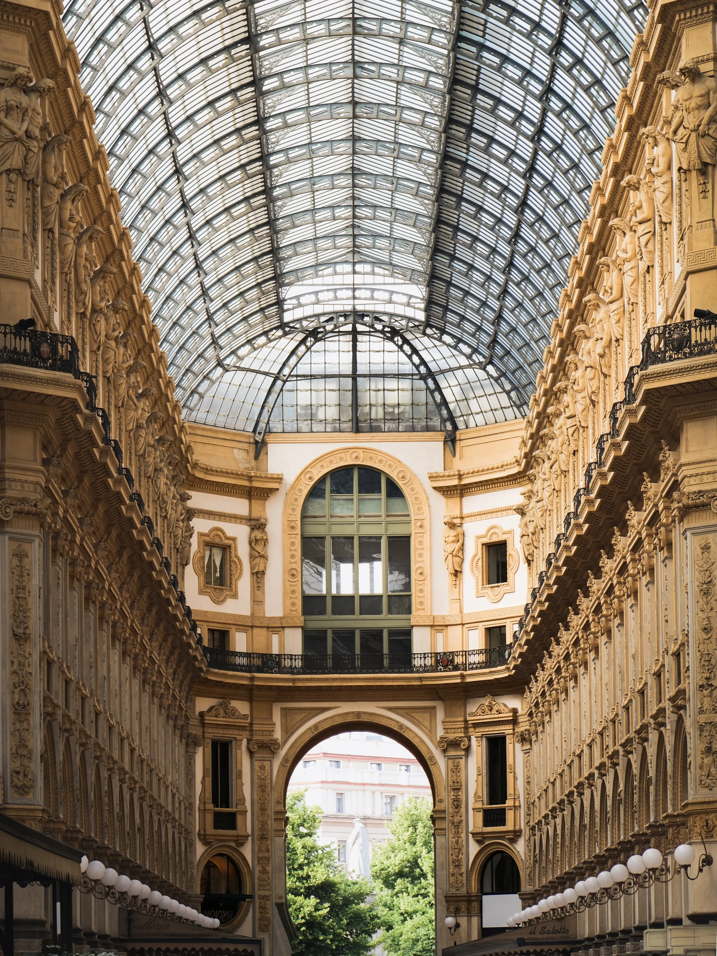 A grand indoor shopping gallery with ornate architecture and a large glass arched ceiling, flanked by detailed stonework and balconies.