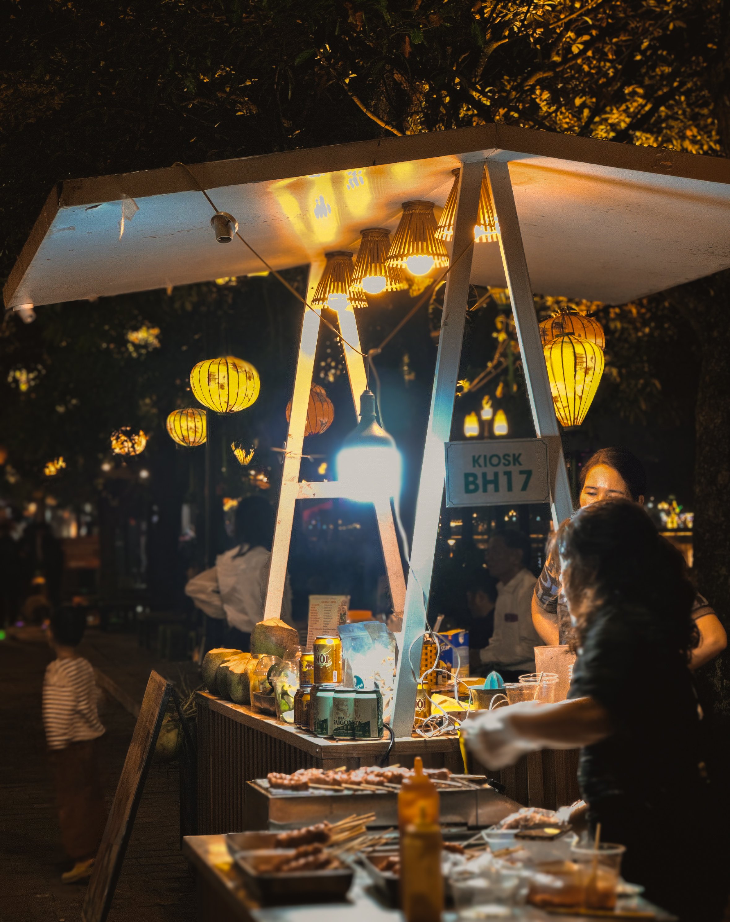 Nighttime scene of a food kiosk with hanging yellow lanterns and string lights, serving skewered grilled food under a illuminated canopy. Several people are attending the stall, with one person preparing food in the foreground.