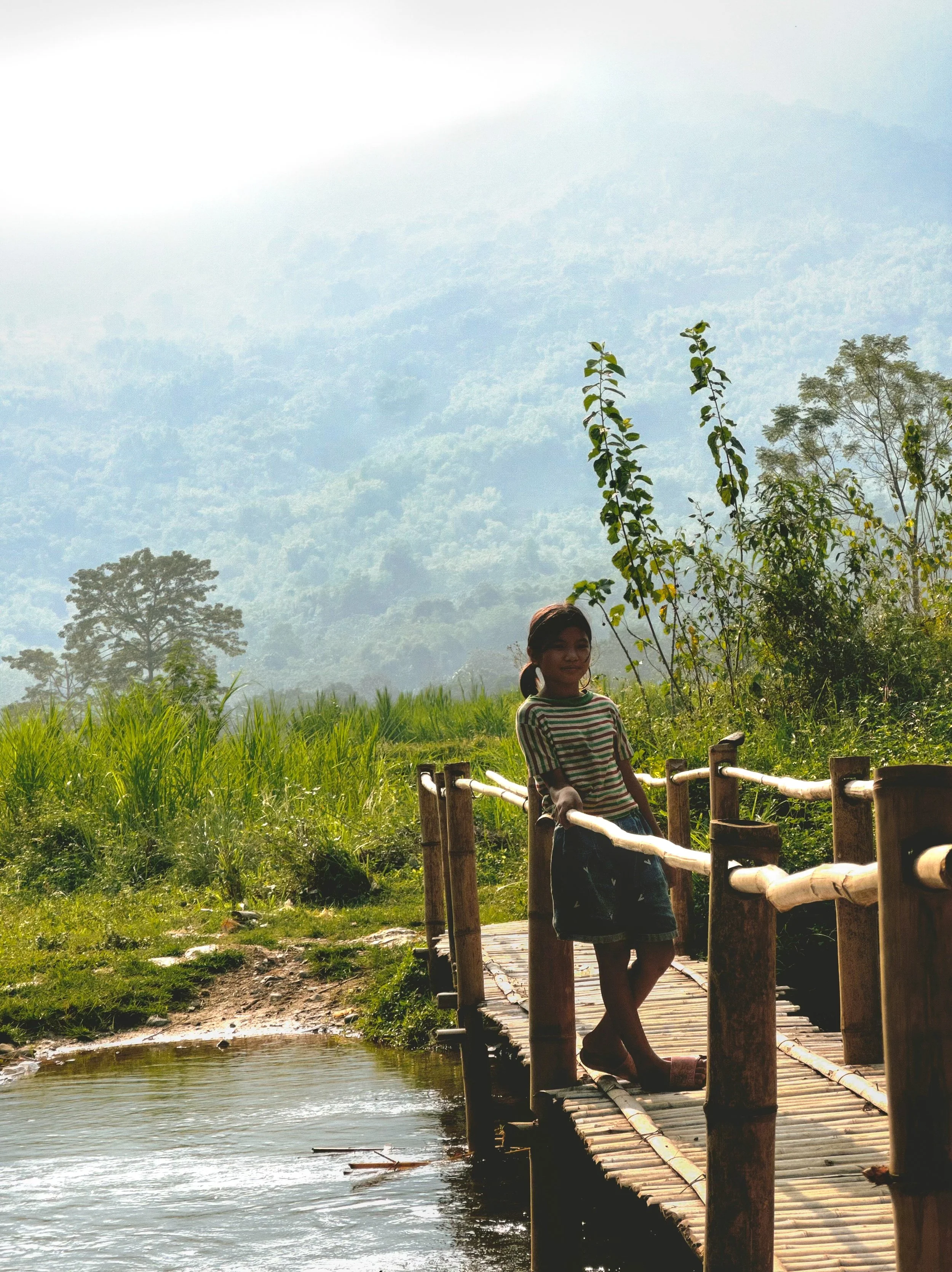 A girl standing on a small wooden bridge over a water body in a green, hilly landscape with trees and mountains in the background.