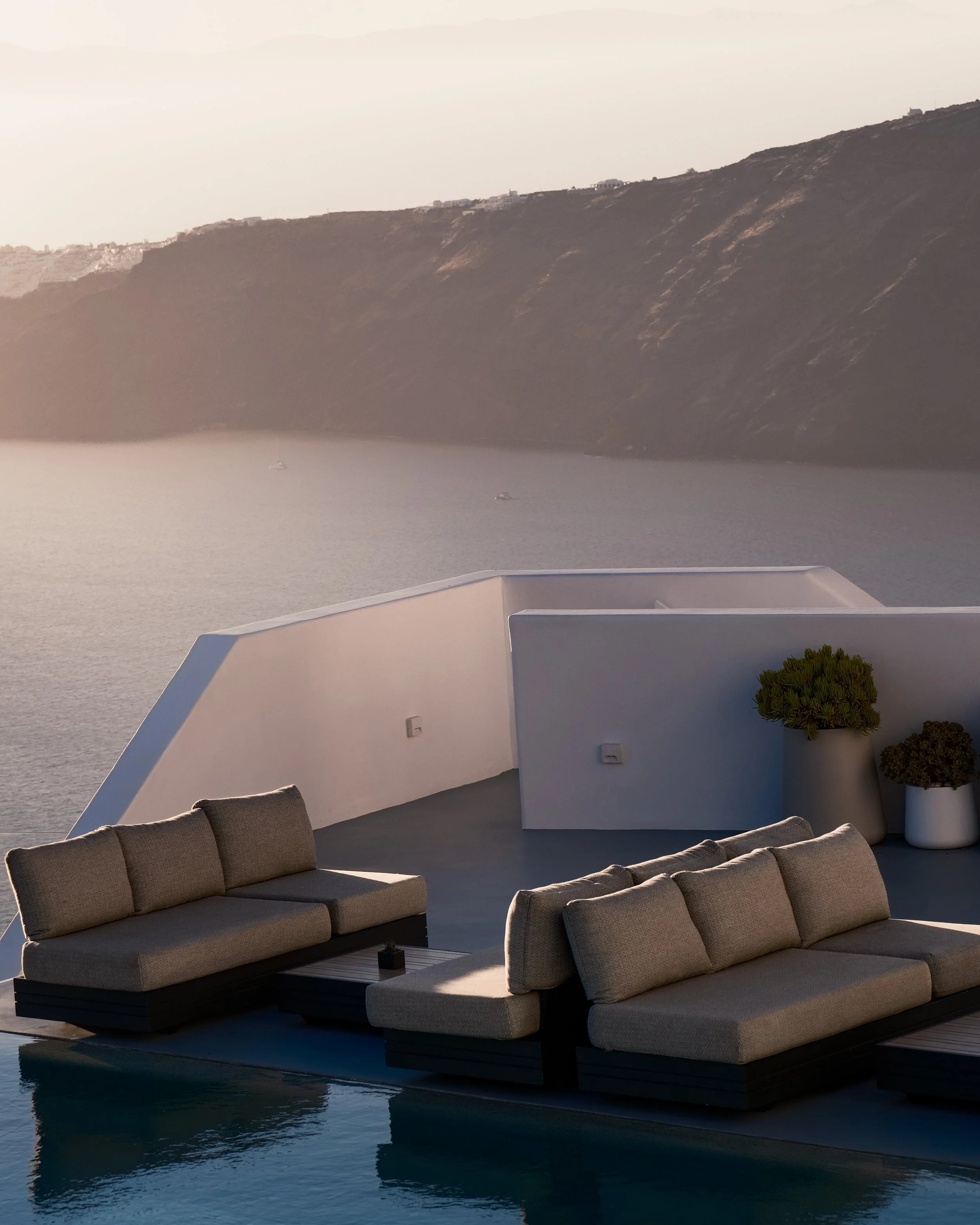 Luxury outdoor seating area with beige cushioned sofa and small table, overlooking water and distant hillside with white buildings.