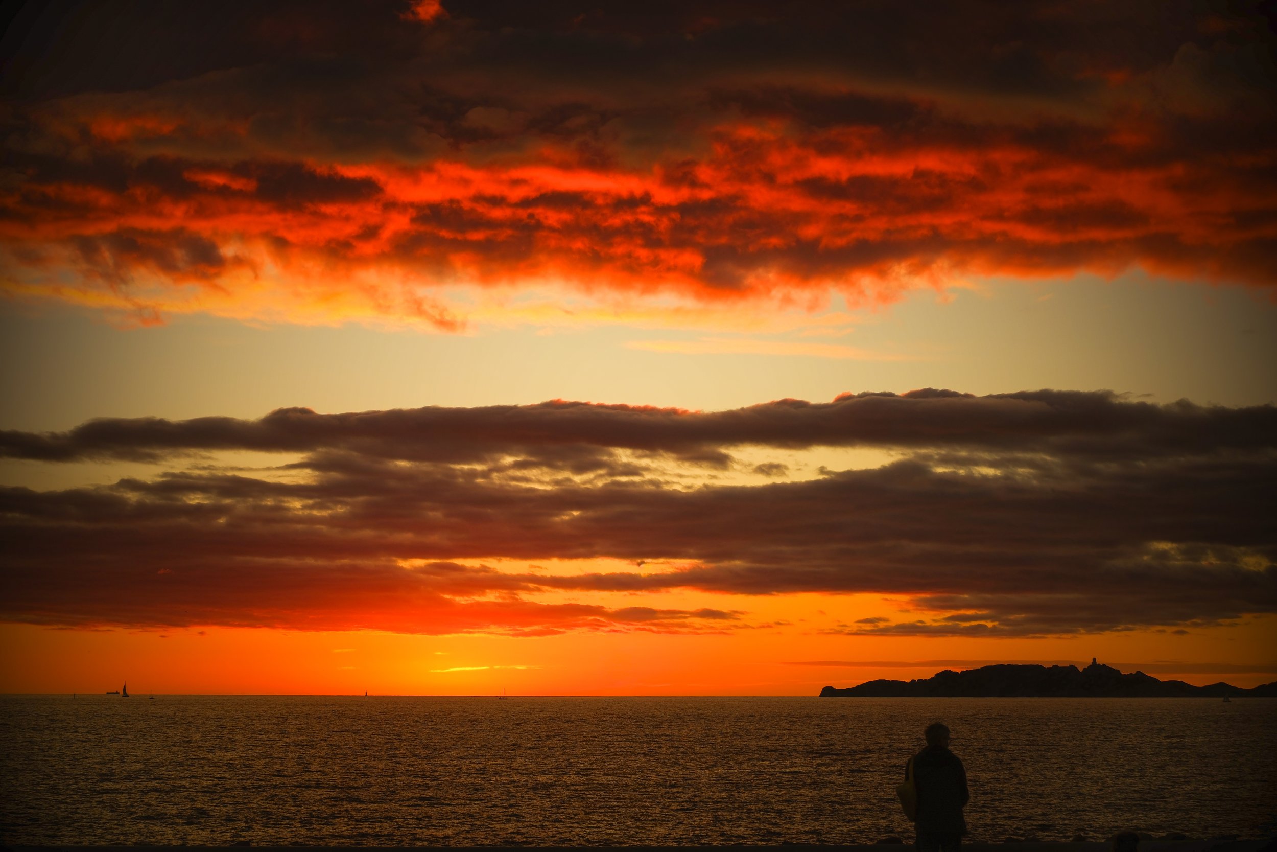 A sunset over the ocean with dark clouds streaked with orange and red, a distant sailboat, and a person standing on the shore