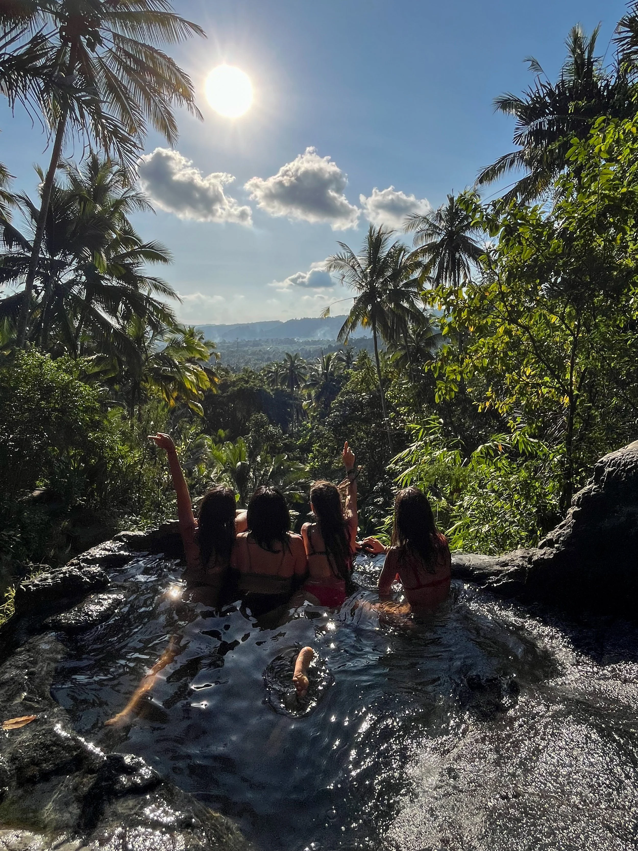 Four women sitting together in a natural pool or hot spring surrounded by lush tropical trees and palm trees under a bright sunny sky, with a view of distant hills.