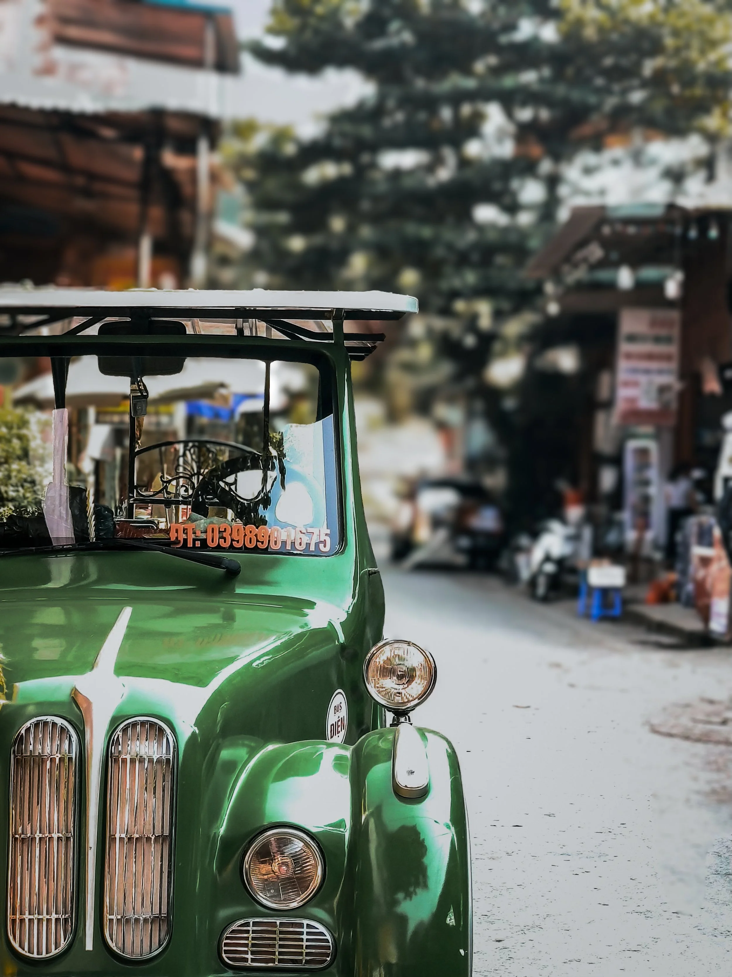 A vintage green tuk-tuk parked on a street with blurred buildings and trees in the background.