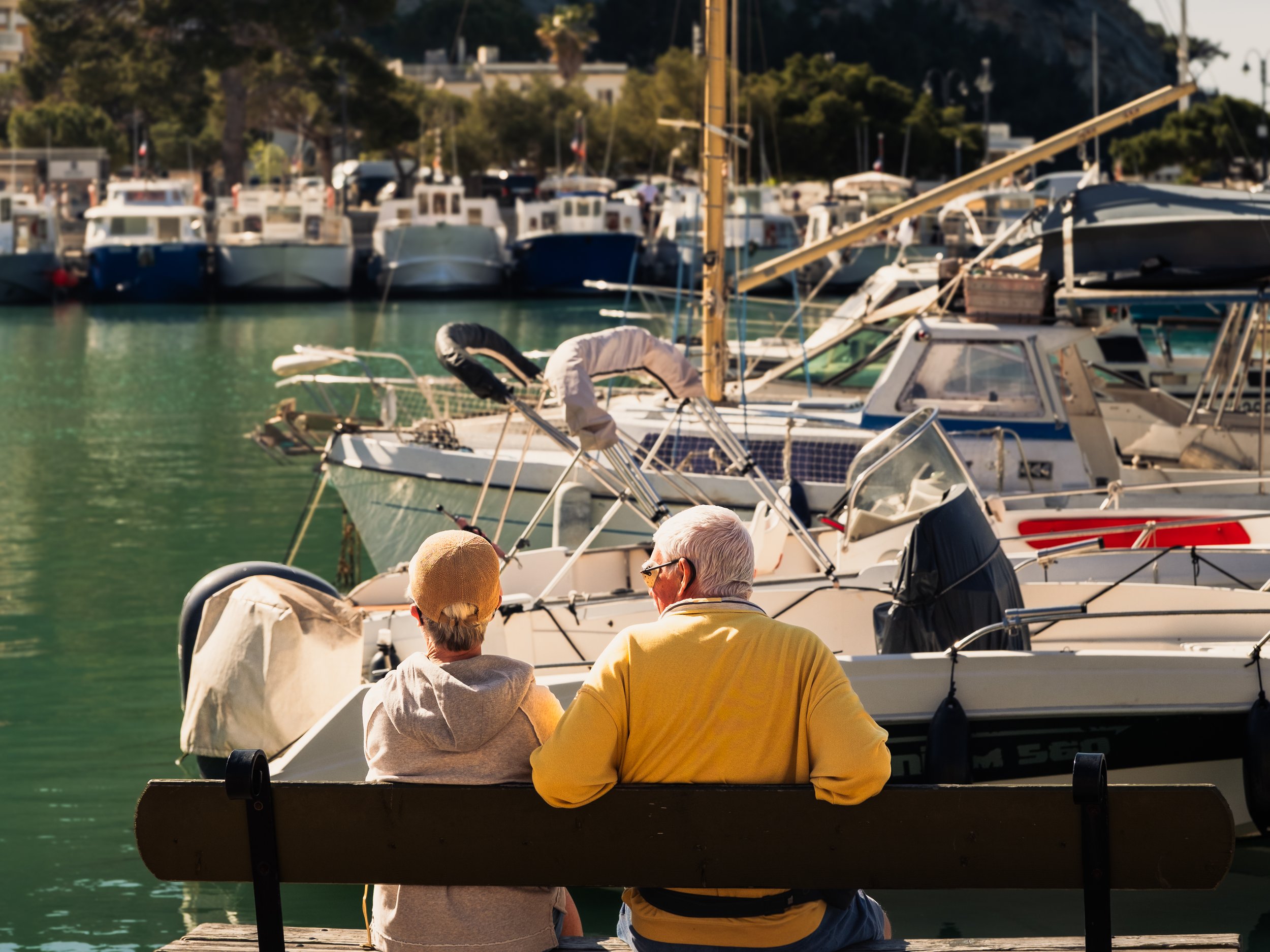 Two men sitting in a boat at a marina, with multiple boats docked on the water behind them, some with protective covers, and gathering trees and buildings in the background.