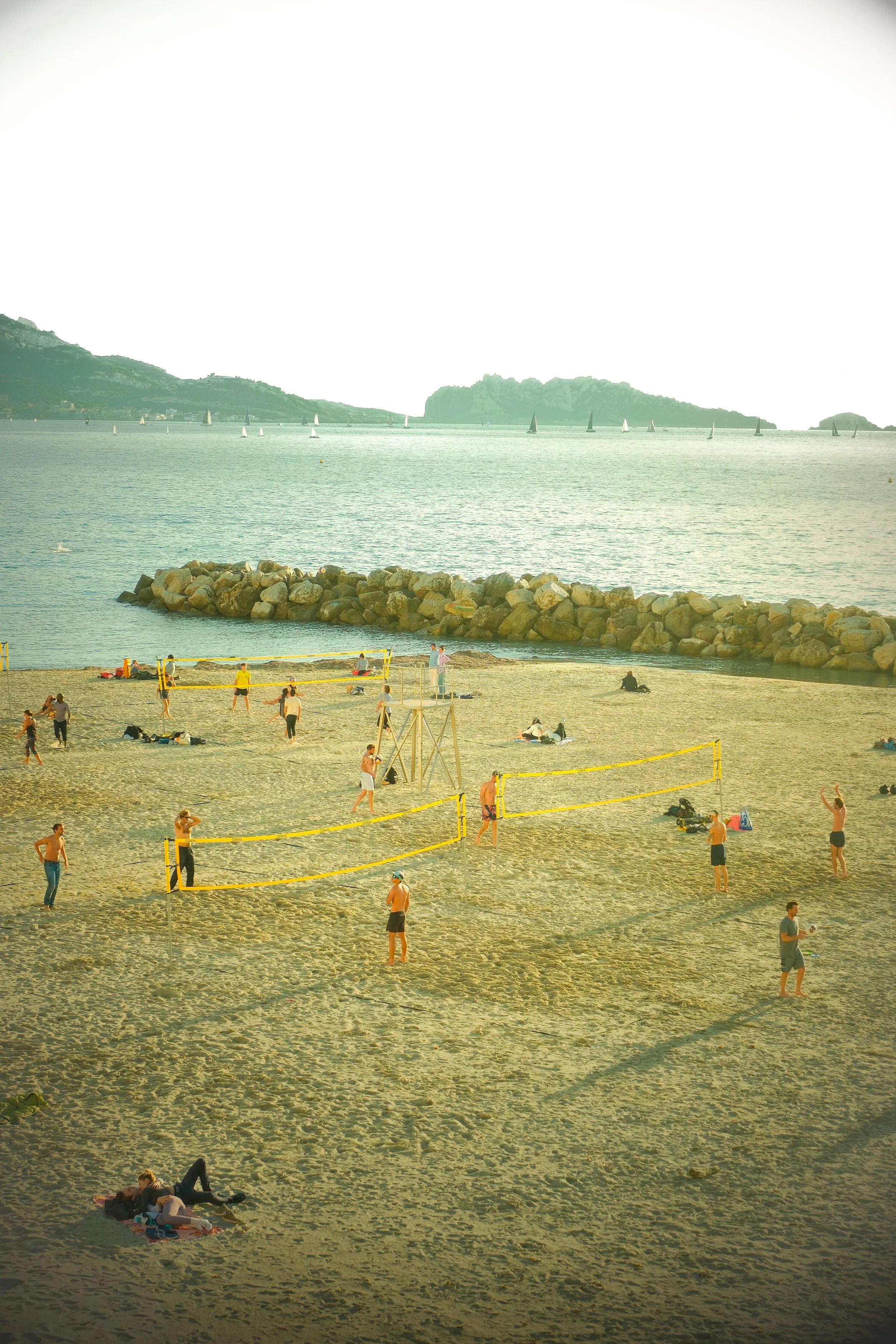 People playing beach volleyball near the shore with sailboats and islands in the background.