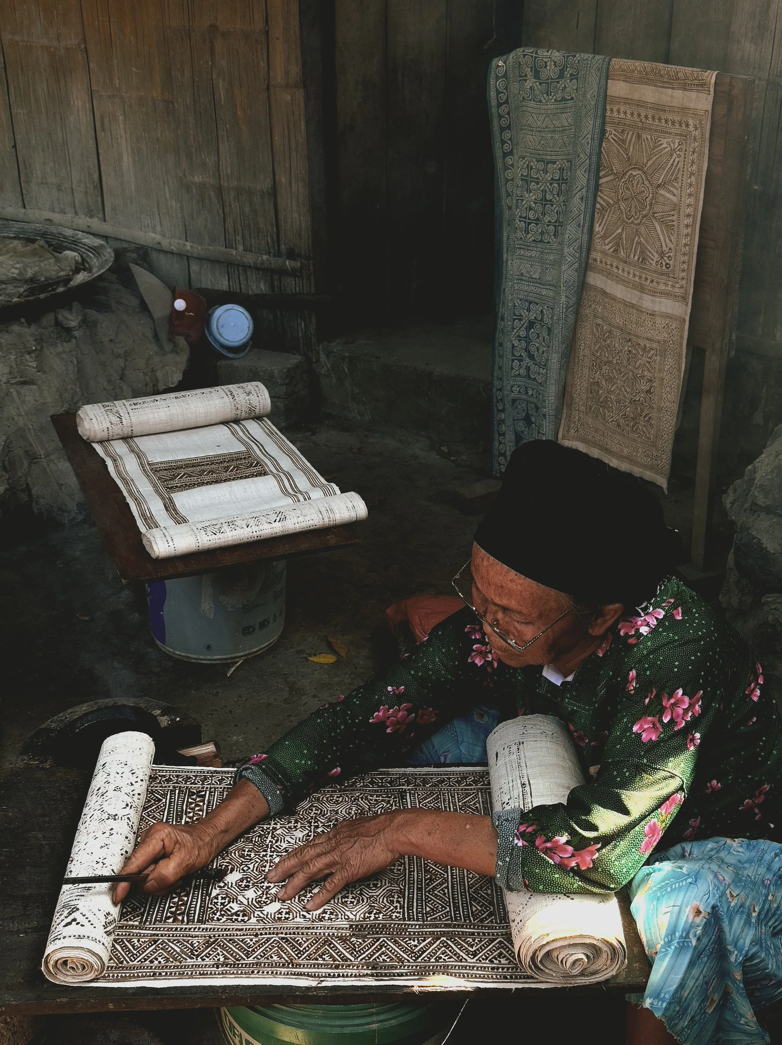 An elderly woman wearing glasses and a black headscarf is sitting on the ground and weaving or embroidering a large piece of fabric with intricate patterns. She is in a rustic room with wooden walls, and textile pieces hang behind her.