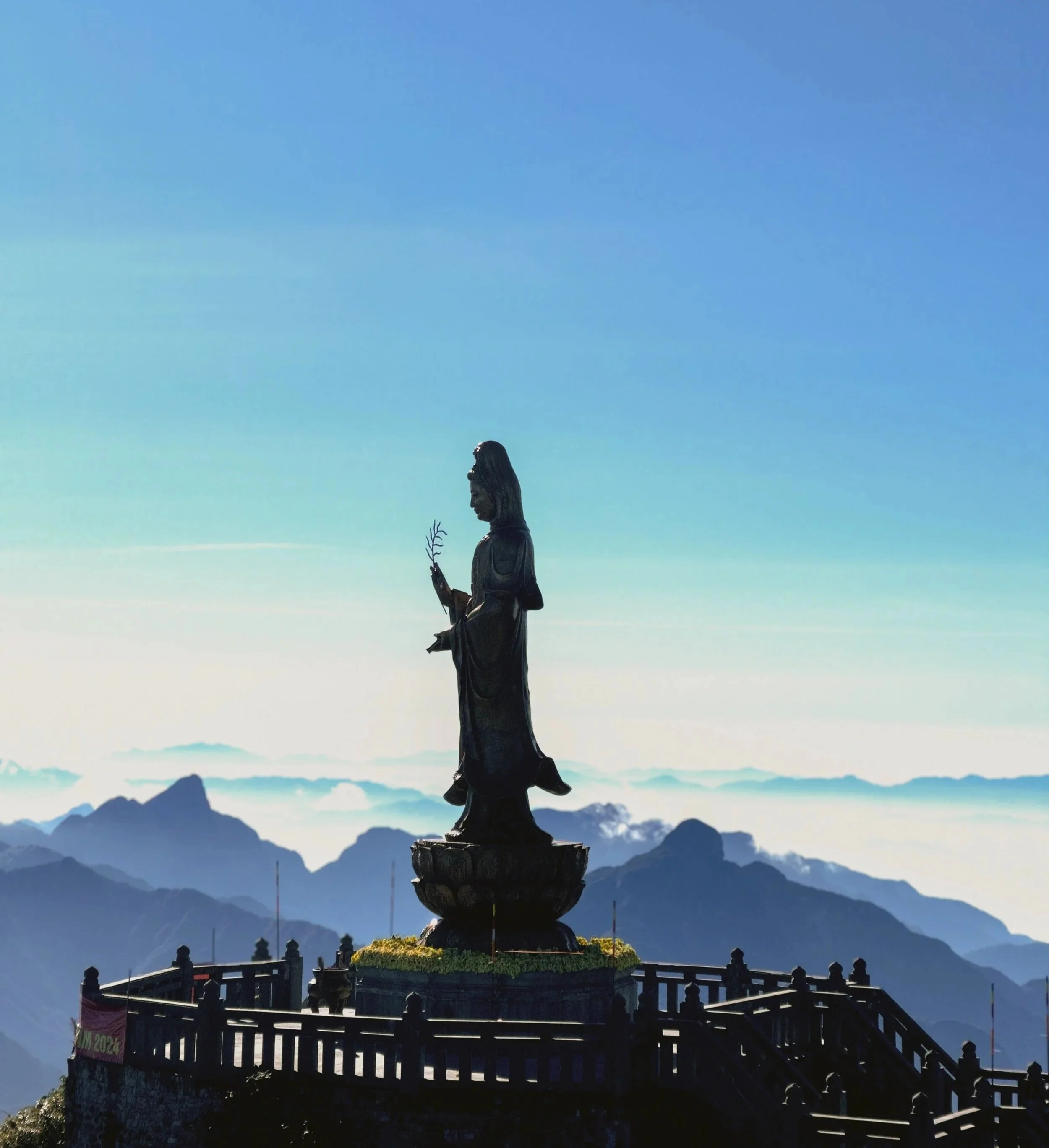 Statue of a woman holding a small tree, standing on a pedestal at a mountain viewpoint against a backdrop of mountain ranges and a blue sky.