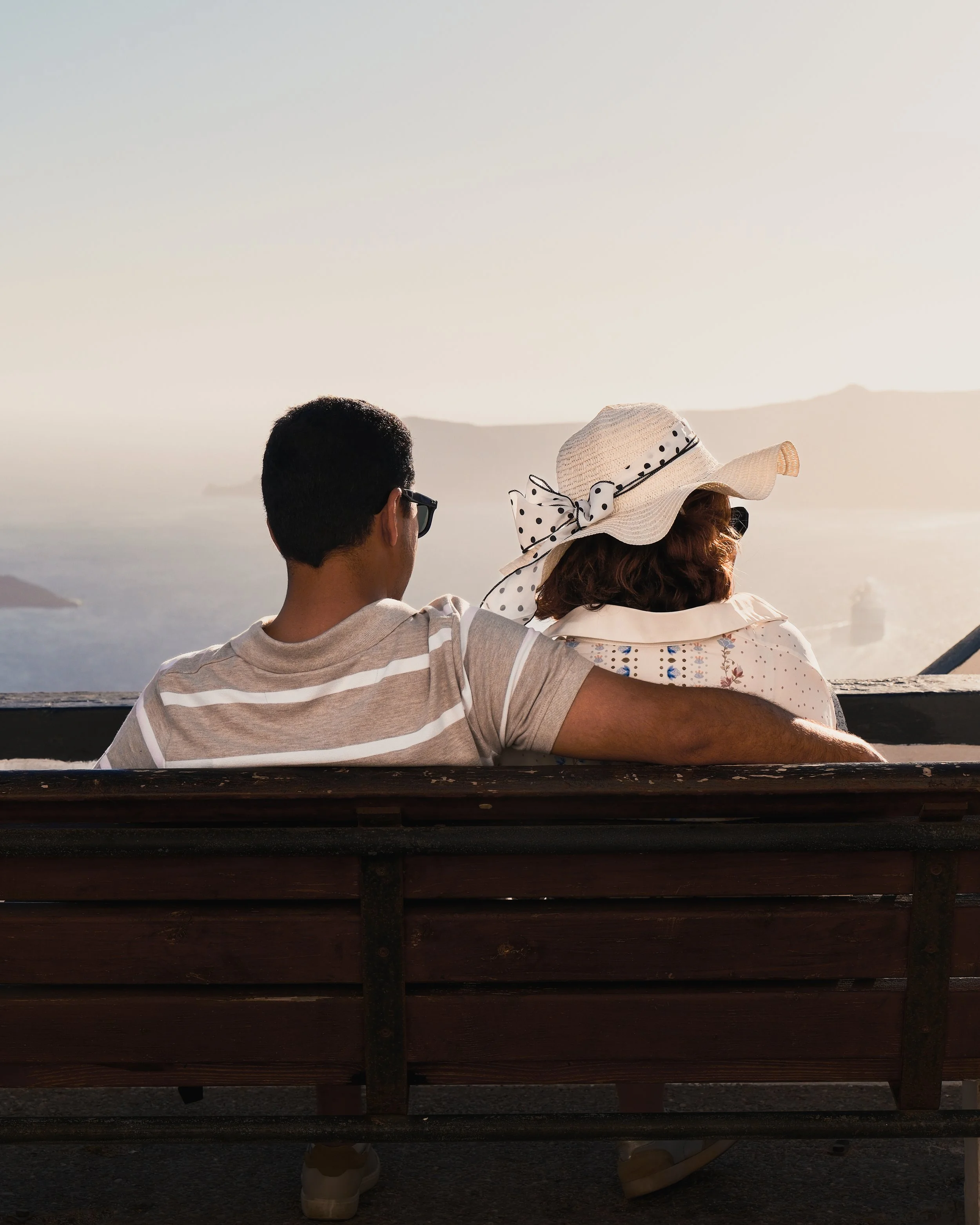 A man and a woman sitting together on a wooden bench, looking out at a body of water and distant mountains, with the man’s arm around the woman, during sunset.