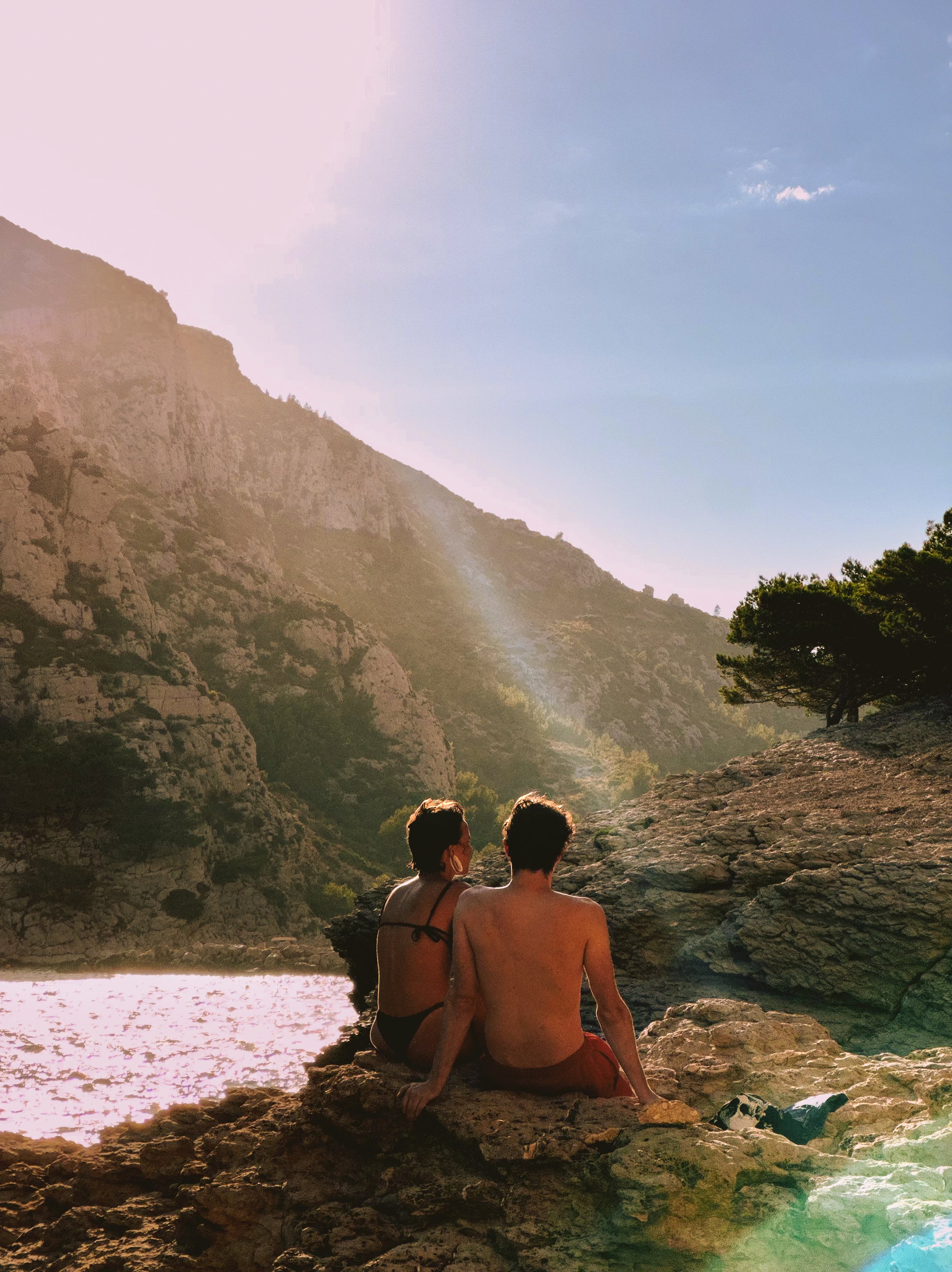 A man and woman sitting on rocks by a river with mountainous landscape and trees in the background, during sunset or sunrise.