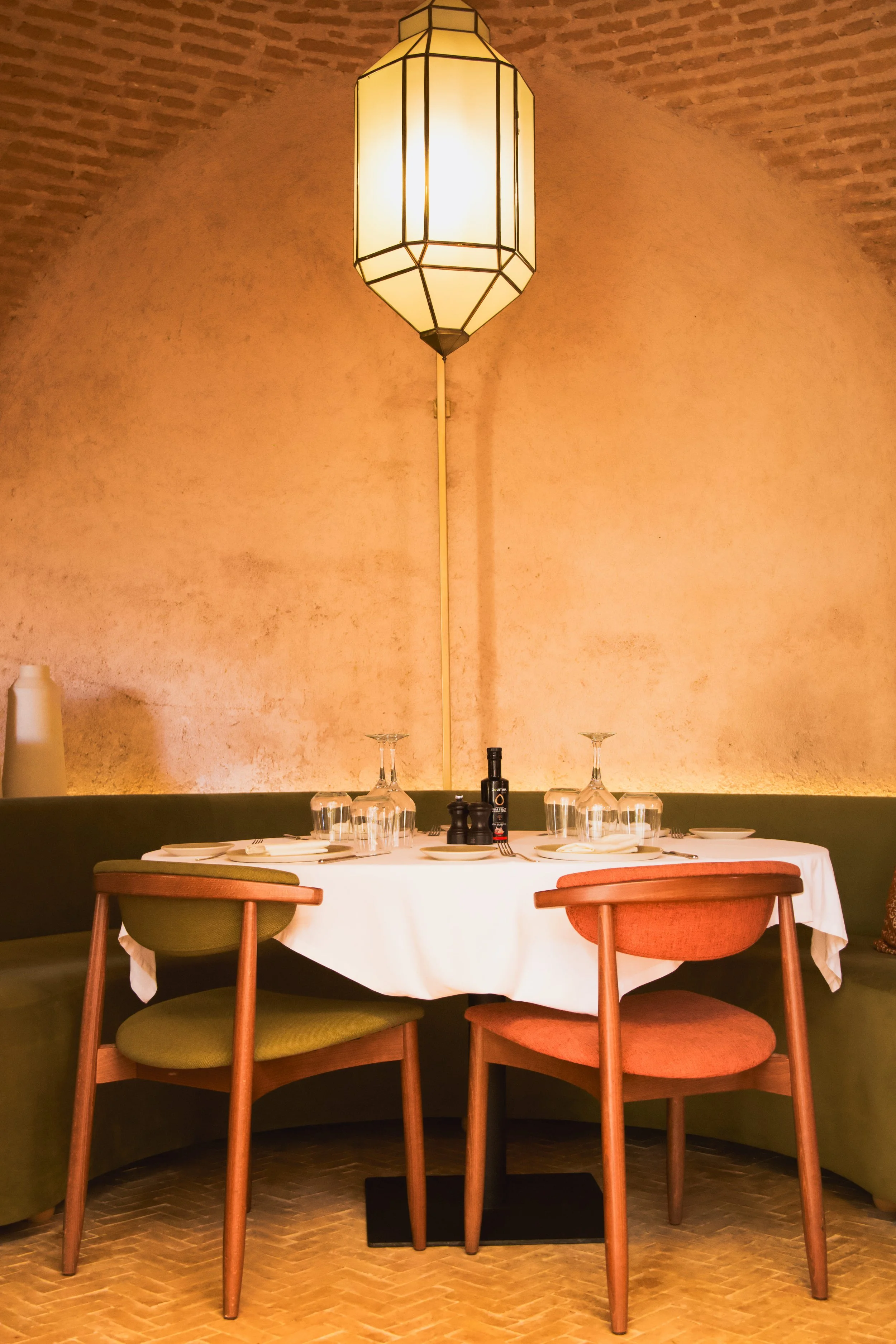 A round dining table set for four with plates, glasses, and silverware in a restaurant with warm lighting, a large geometric pendant light, and a textured peach-colored wall.