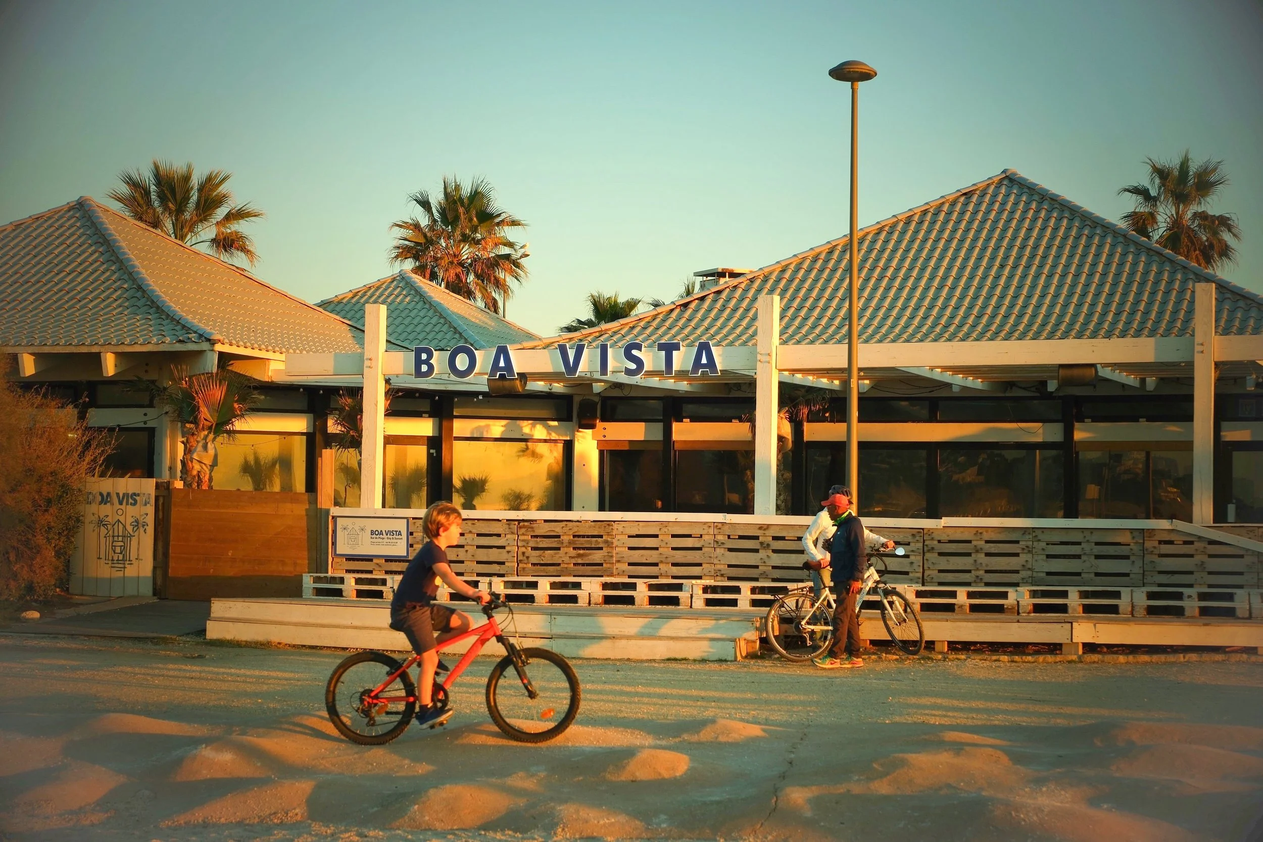 People with bicycles near the Boa Vista restaurant with palm trees in the background at sunset.