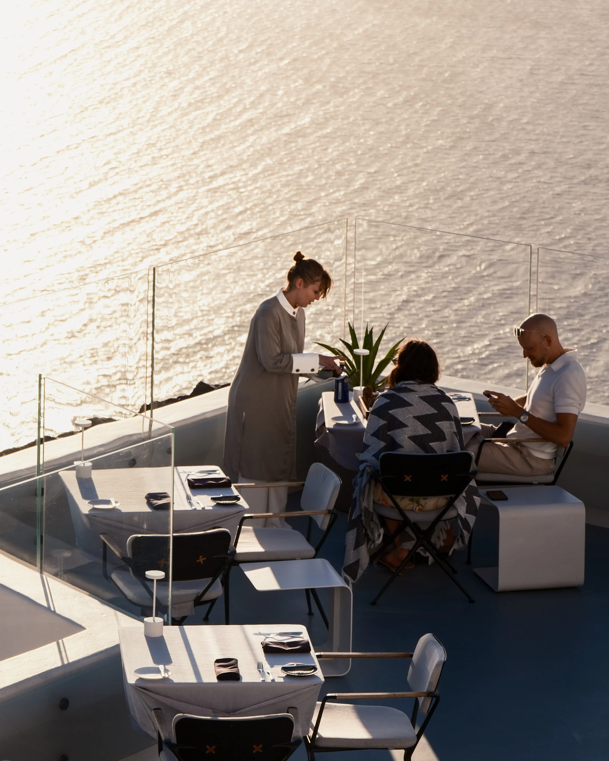 A group of people dining tables on a terrace overlooking a body of water during sunset, with the woman wearing a gray coat focusing on her phone.