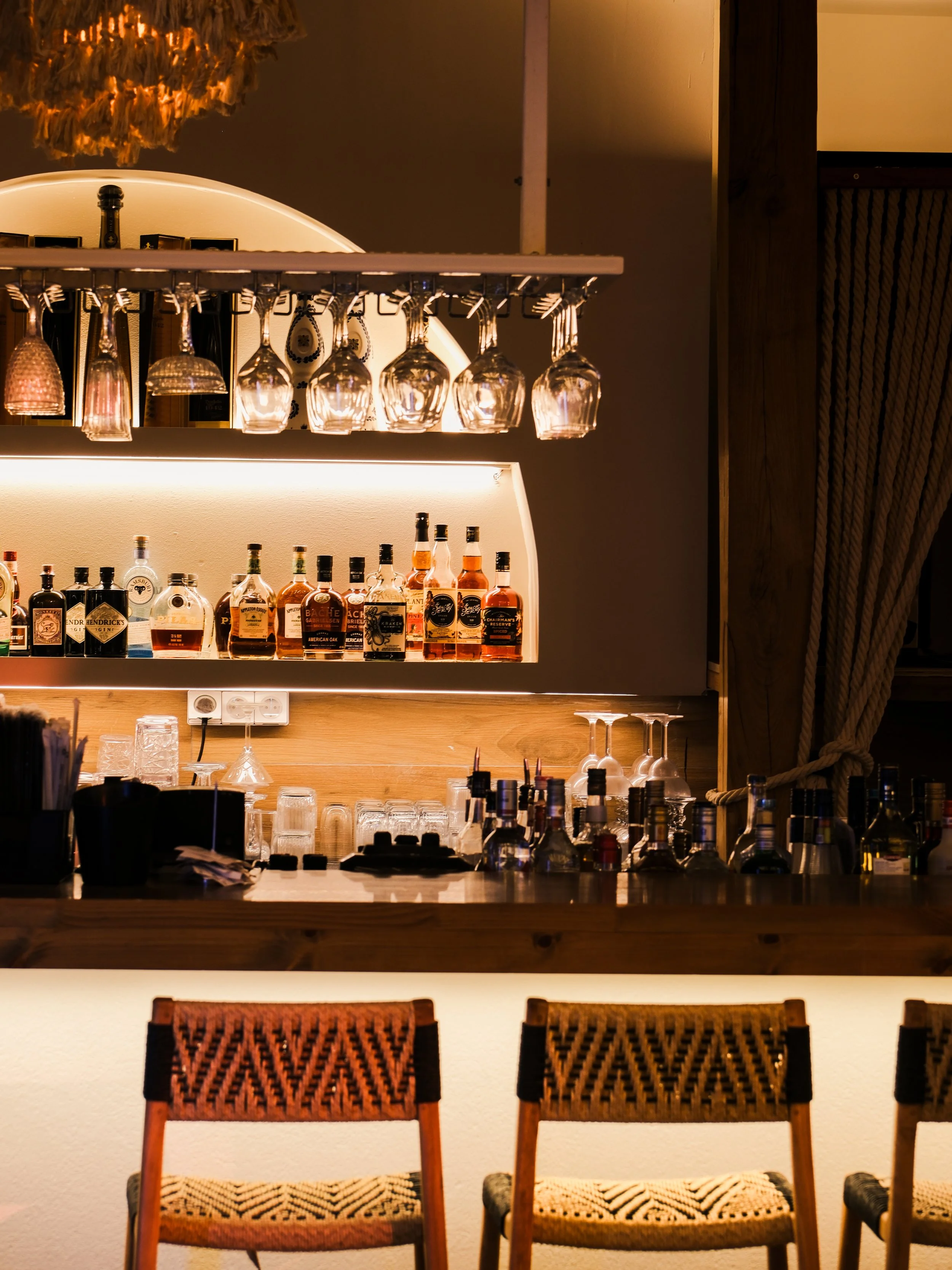 Bar with liquor bottles, wine glasses hanging upside down, and chairs in the foreground, warm lighting creating a cozy ambiance.