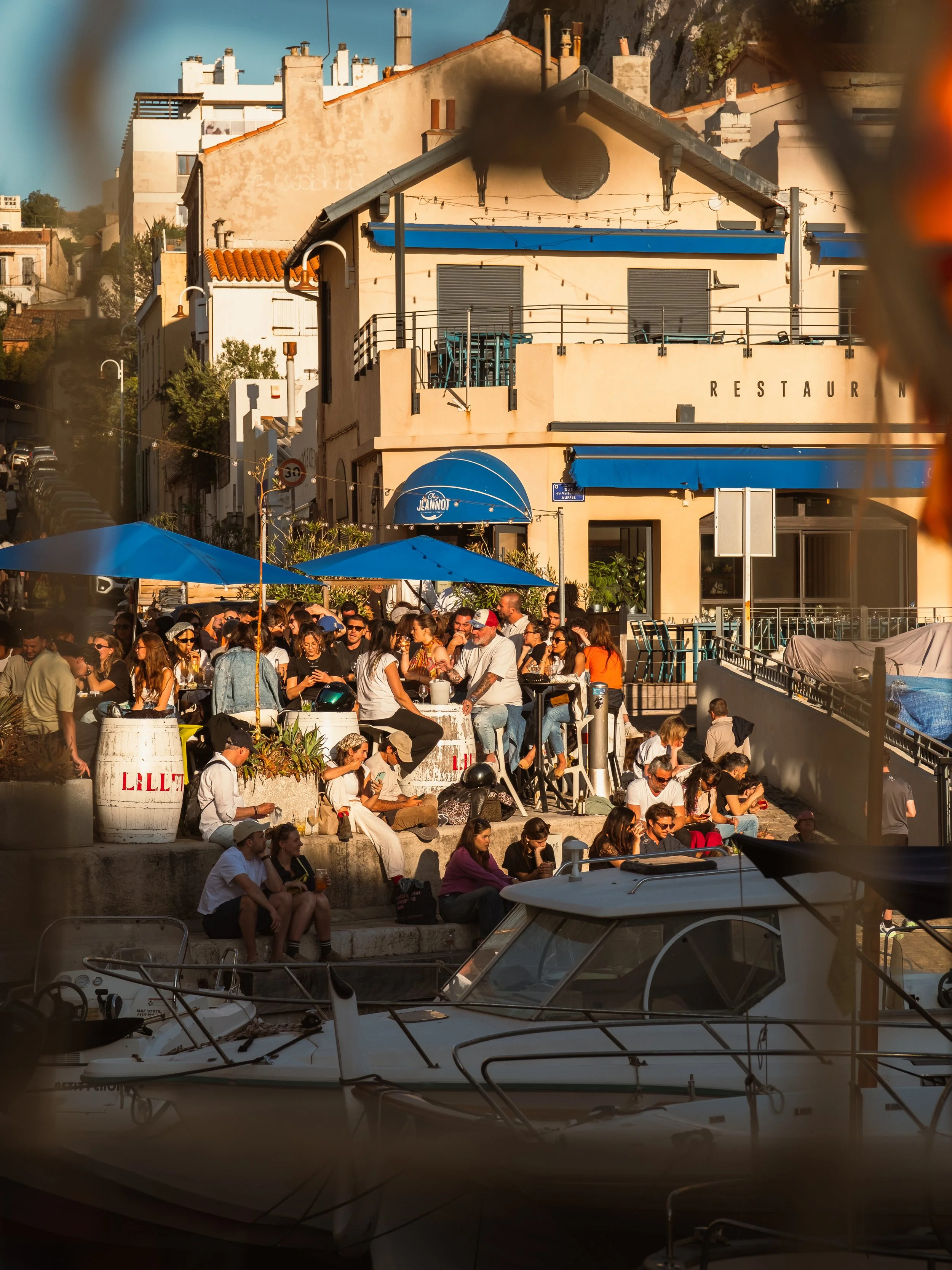 Crowd of people sitting and standing at an outdoor restaurant or café along a waterfront, with boats docked in the foreground and buildings with terracotta roofs in the background.