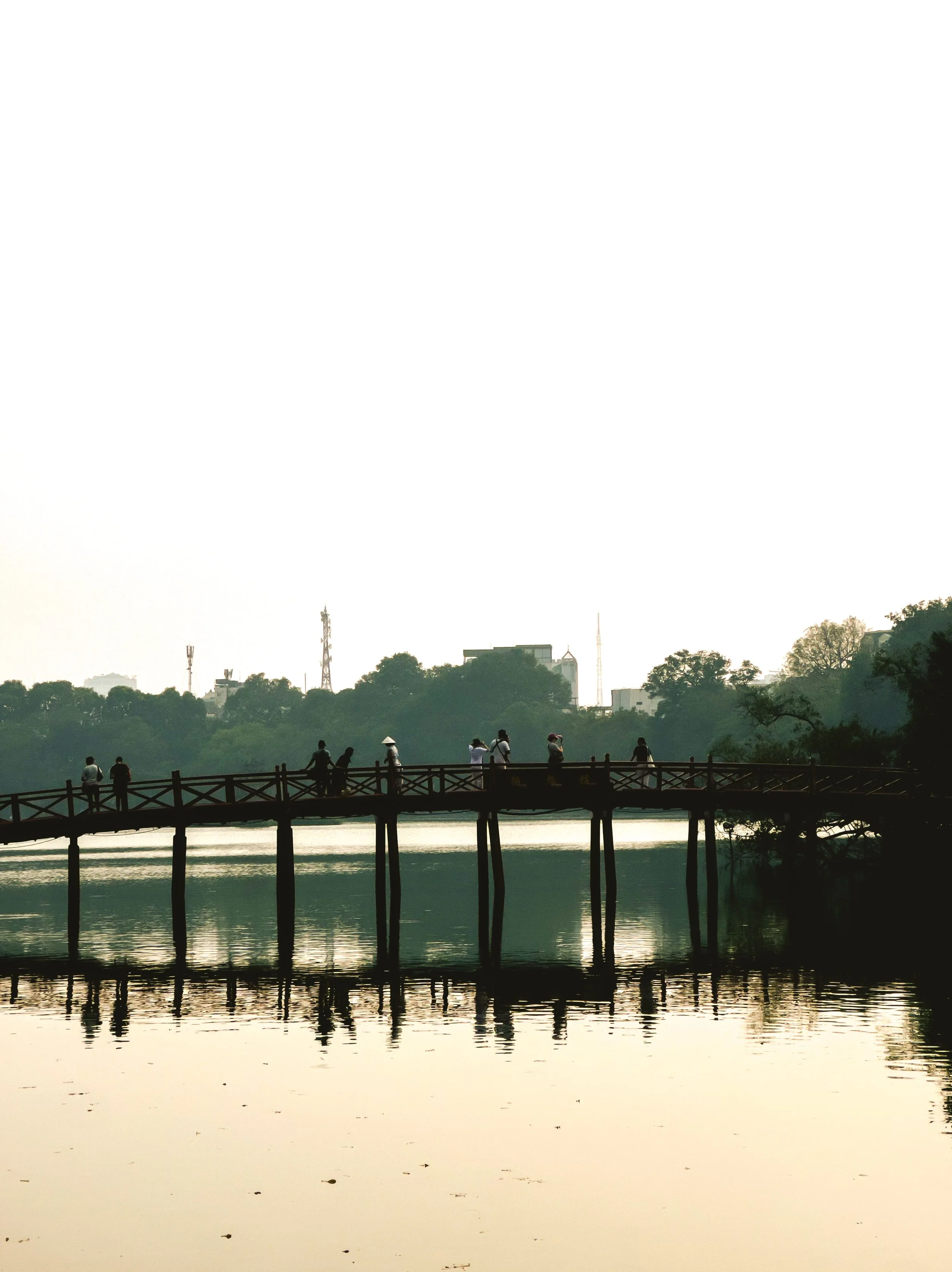 People walking across a wooden footbridge over a calm body of water with trees and buildings in the background.