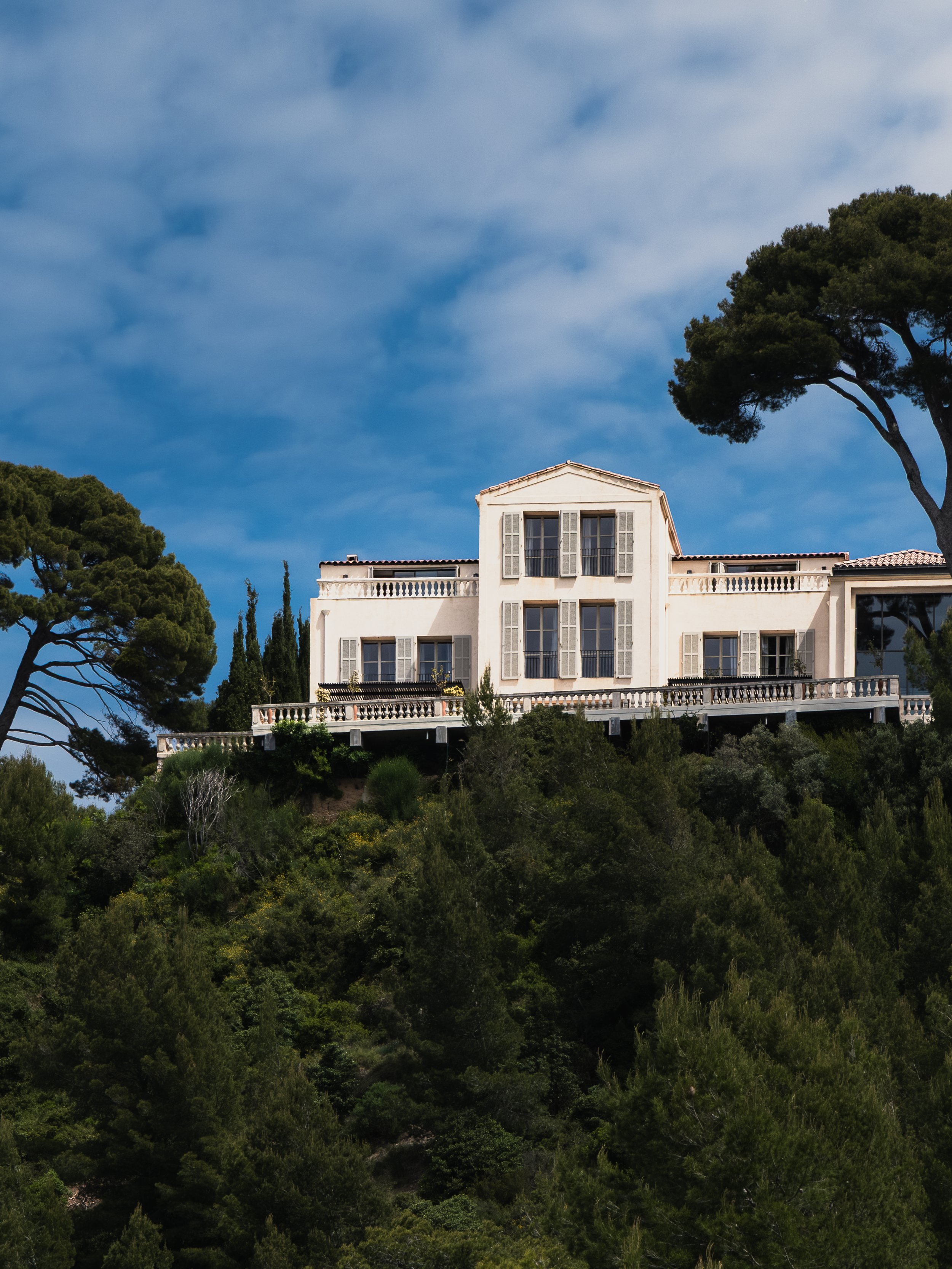 A white house with multiple windows and shutters is perched on a hillside surrounded by green trees and bushes, with a partly cloudy blue sky overhead.