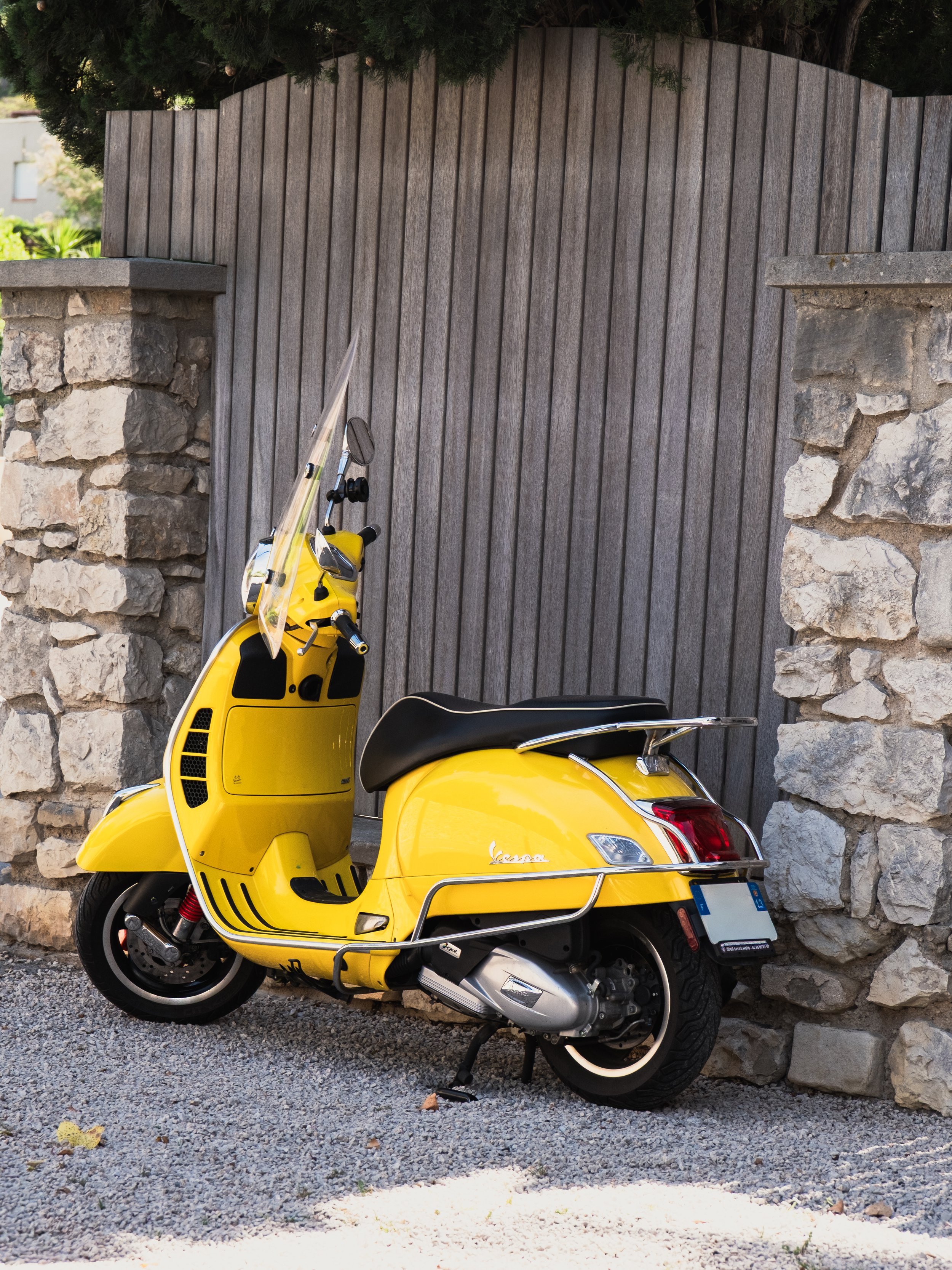 A yellow Vespa scooter parked against a stone wall with a wooden gate in the background.