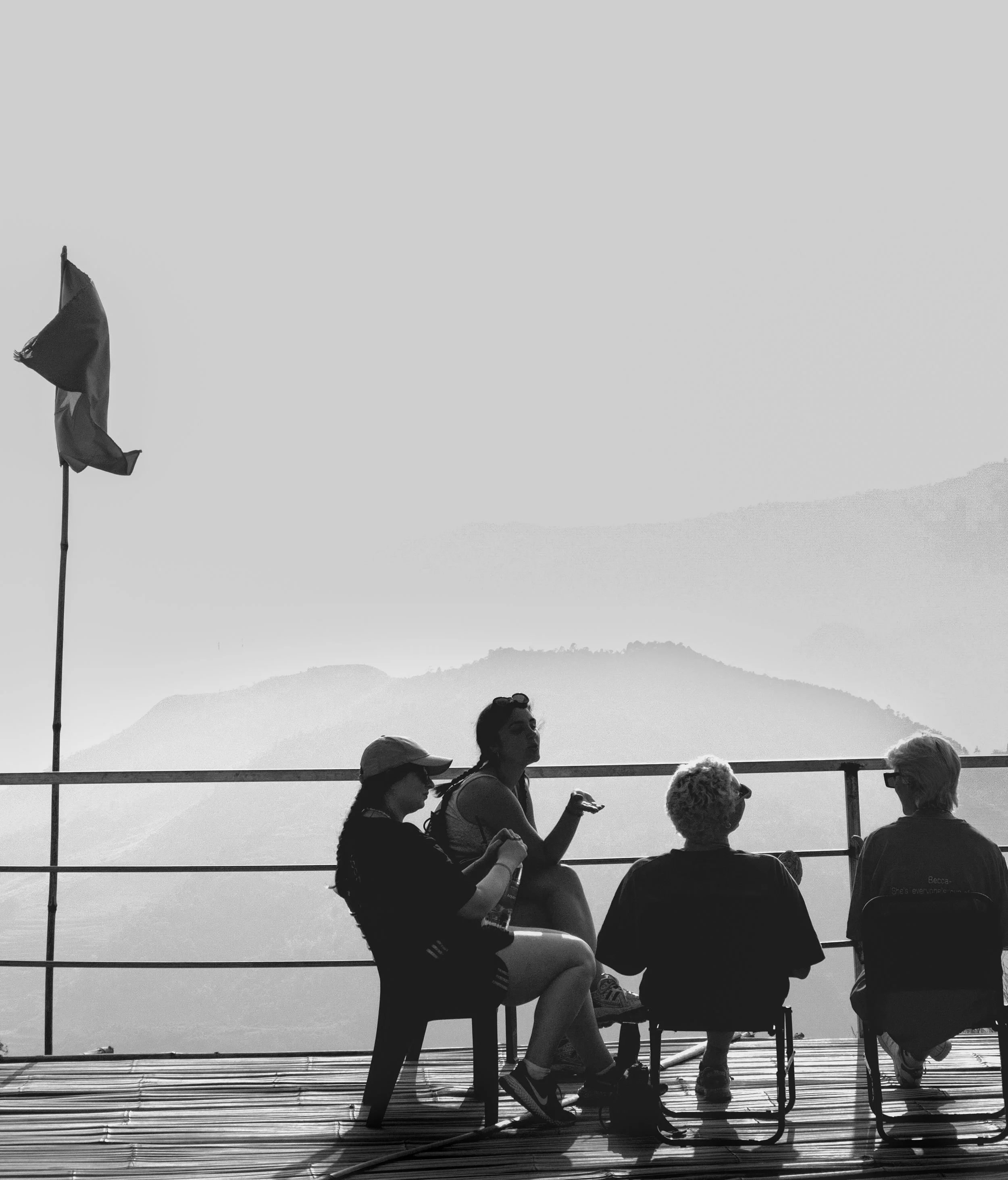 Four women sitting and standing on a deck overlooking mountains, engaged in conversation, with a flag on a pole to the left.