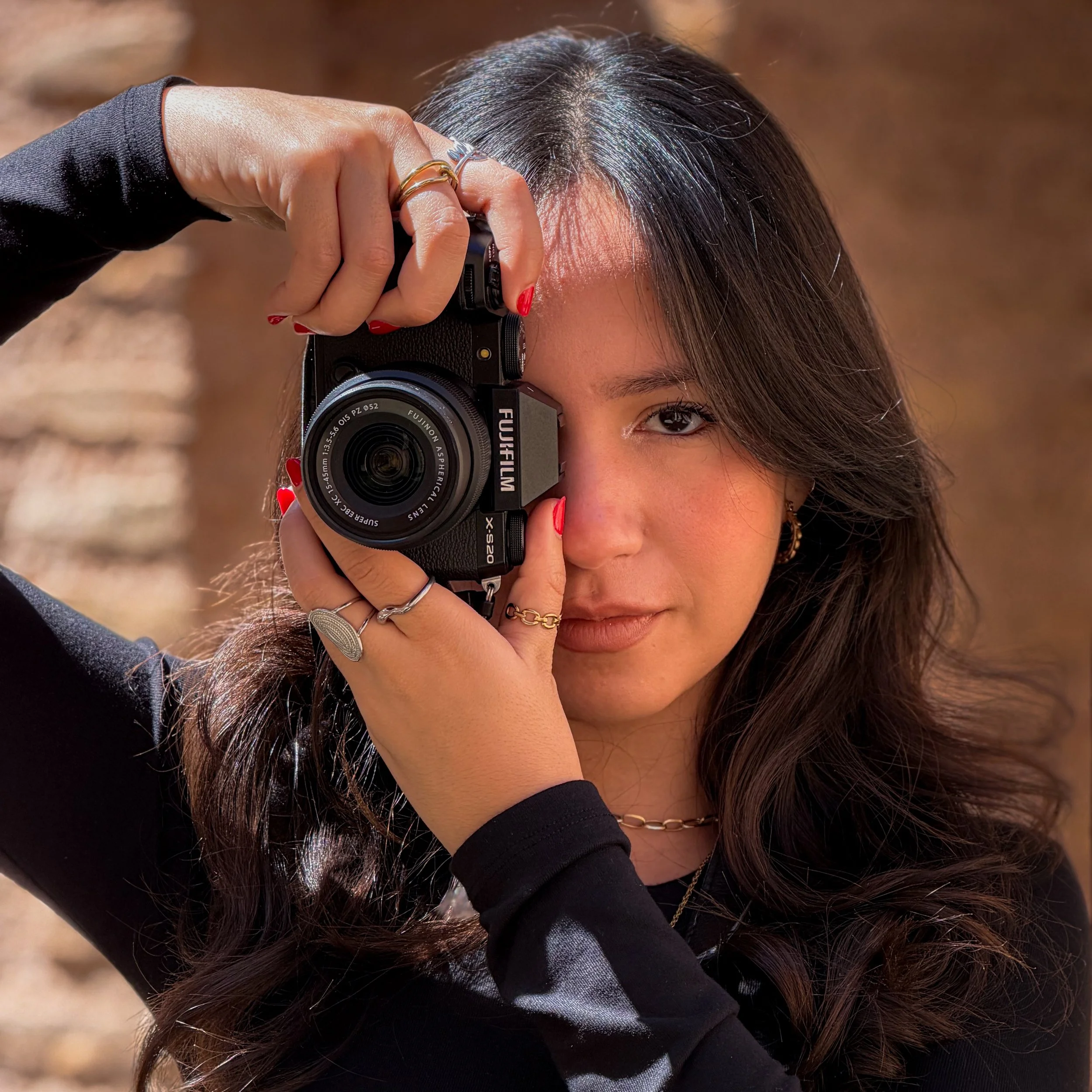 A woman with dark hair and fair skin holding a camera up to her eye, wearing rings and painted nails, outdoors in natural light.