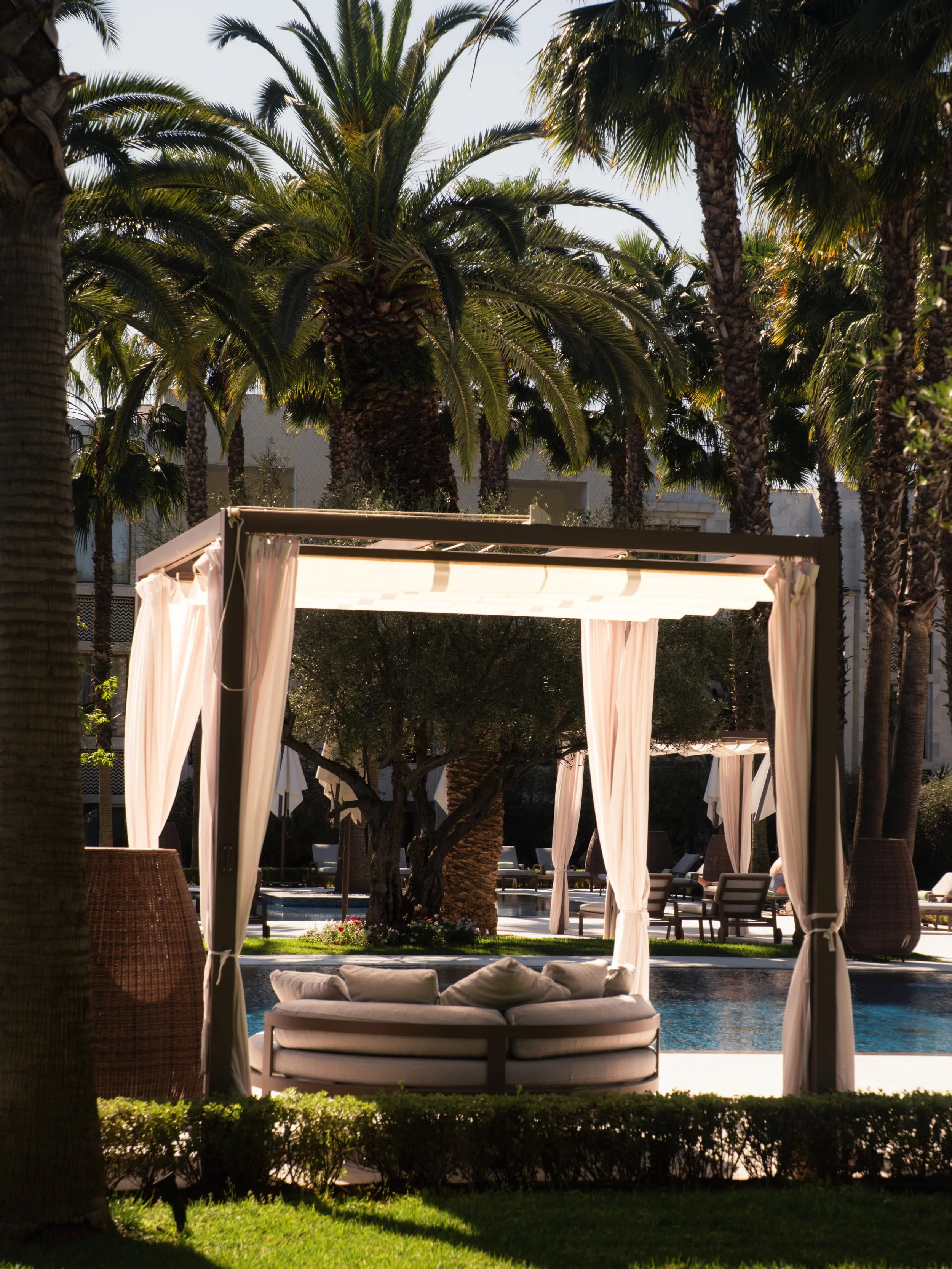 Luxury outdoor pool area with cabanas, lounge chairs, lush palm trees, and a poolside bed with cushions, during daylight.