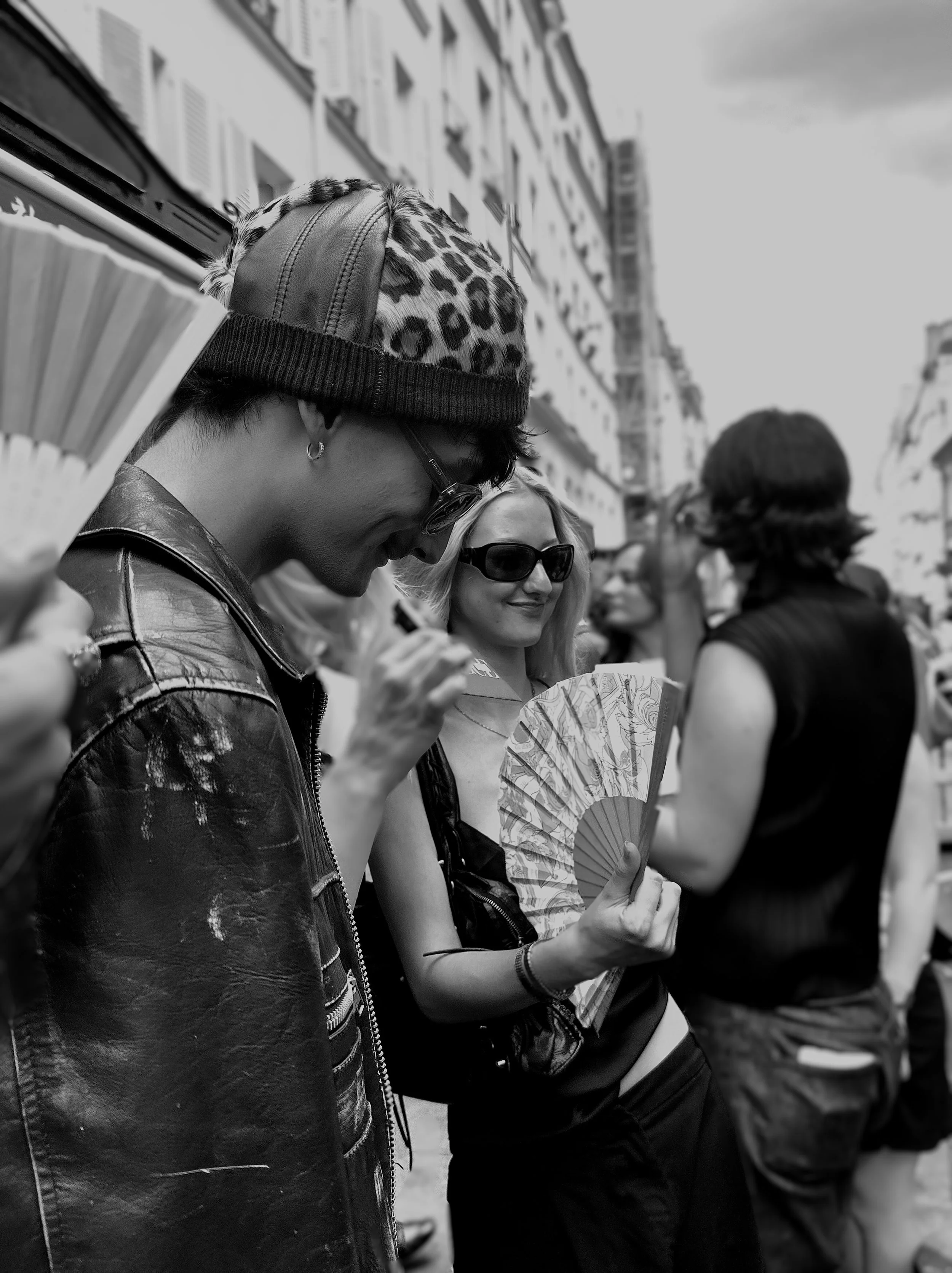People standing outside on a city street, wearing sunglasses, with two women in the center smiling and holding a fan, one wearing a leopard print hat and leather jacket, the other wearing a sleeveless top and a headband.