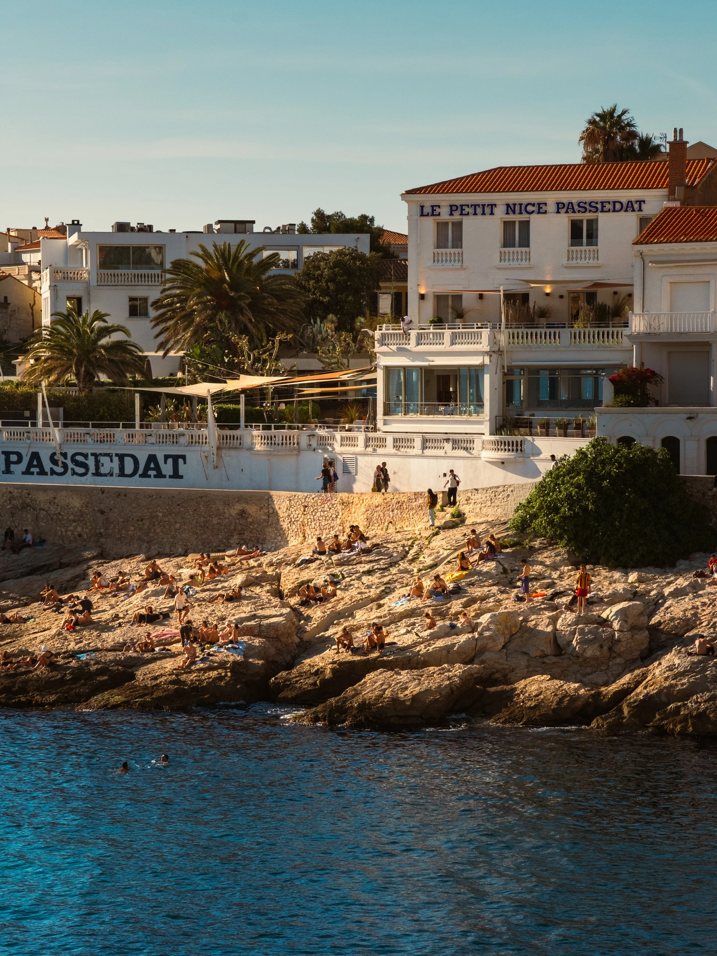 People sunbathing and swimming on rocky shoreline in front of white buildings with palm trees and clear sky.