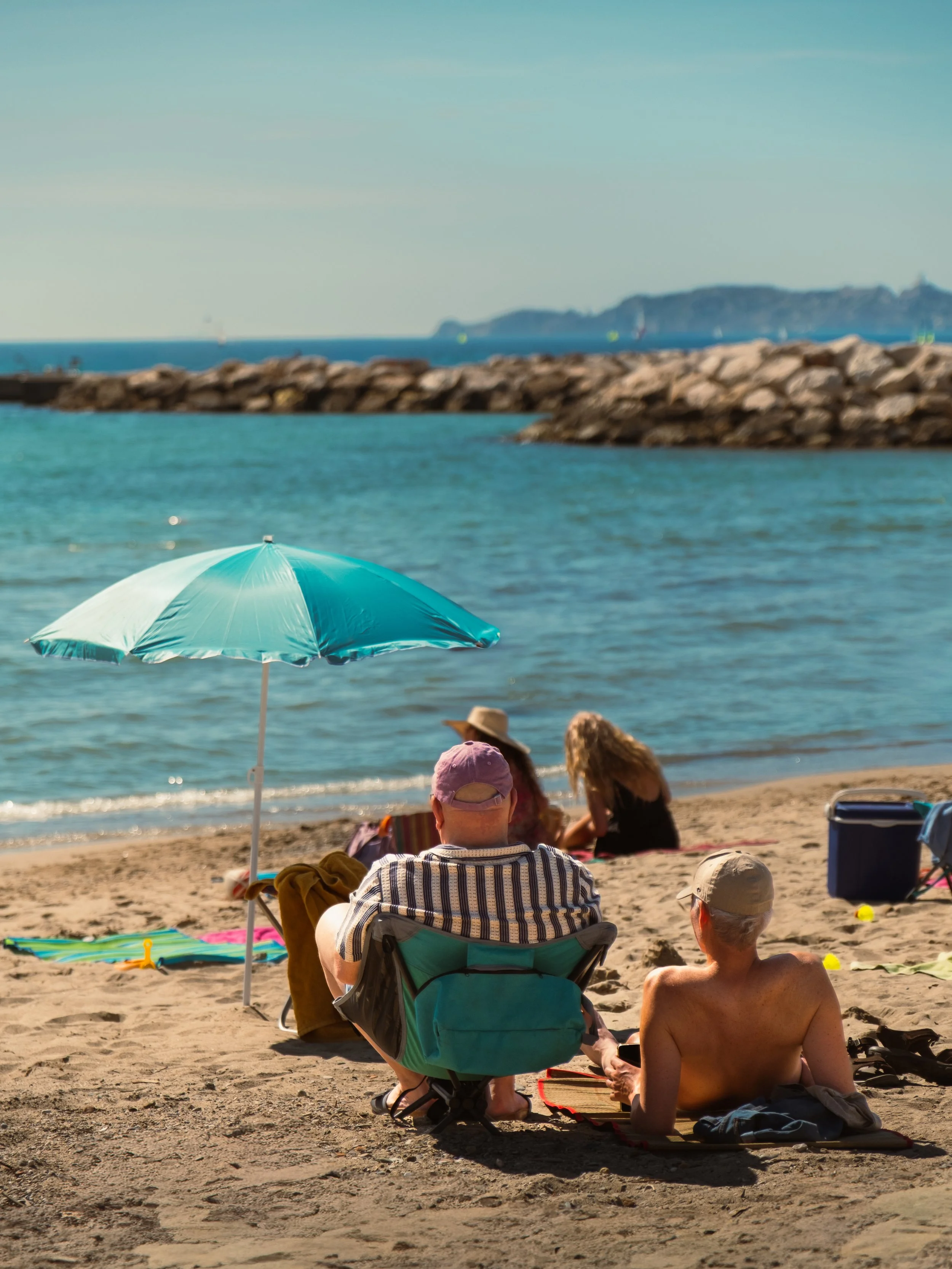 People relaxing on a sandy beach under umbrellas, with the ocean and rocky breakwater in the background.