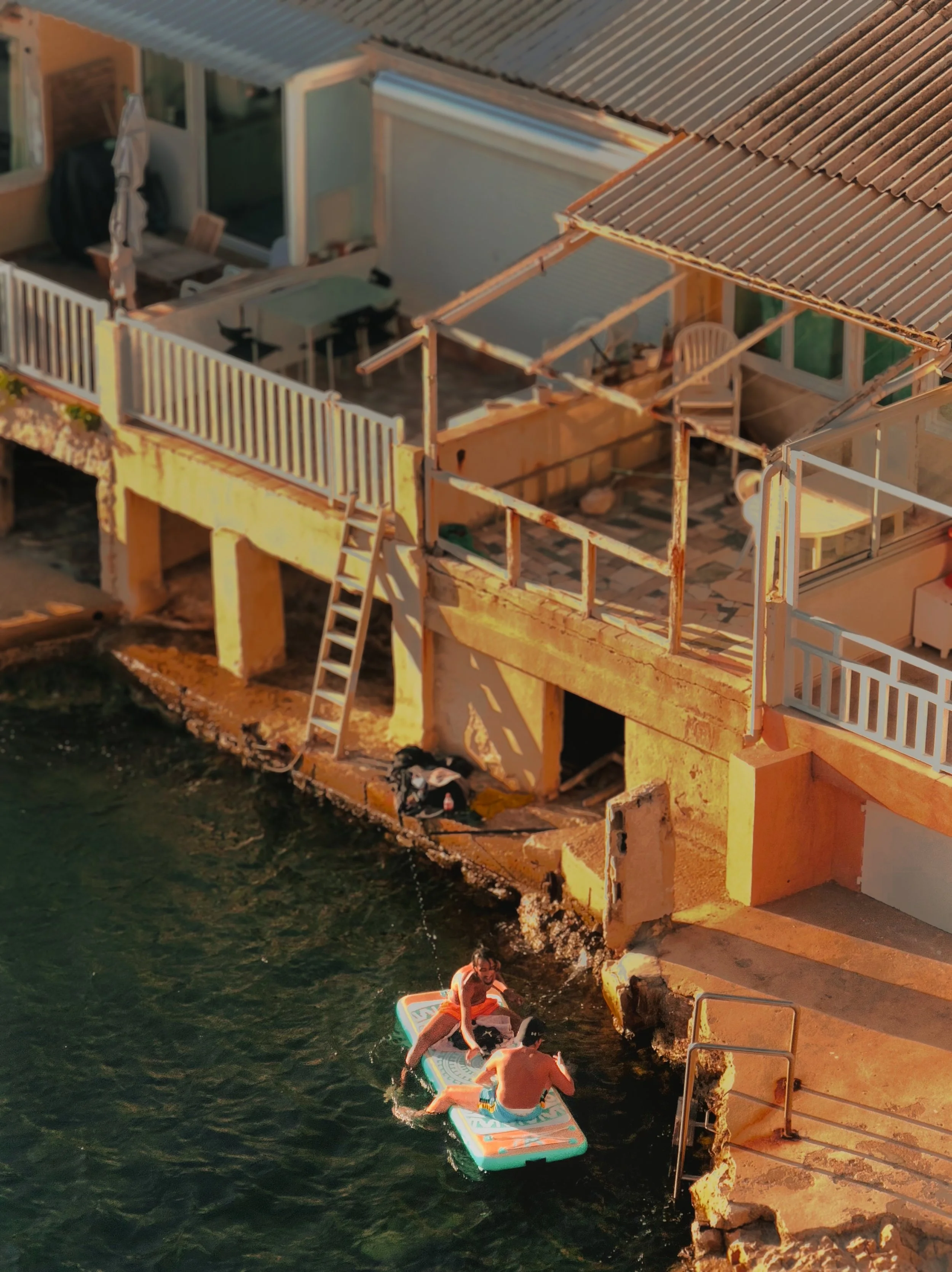 Two people sitting on a paddleboard near a cliffside house by the water, with a ladder leading from the house to the water, and the house having a patio area with outdoor furniture.