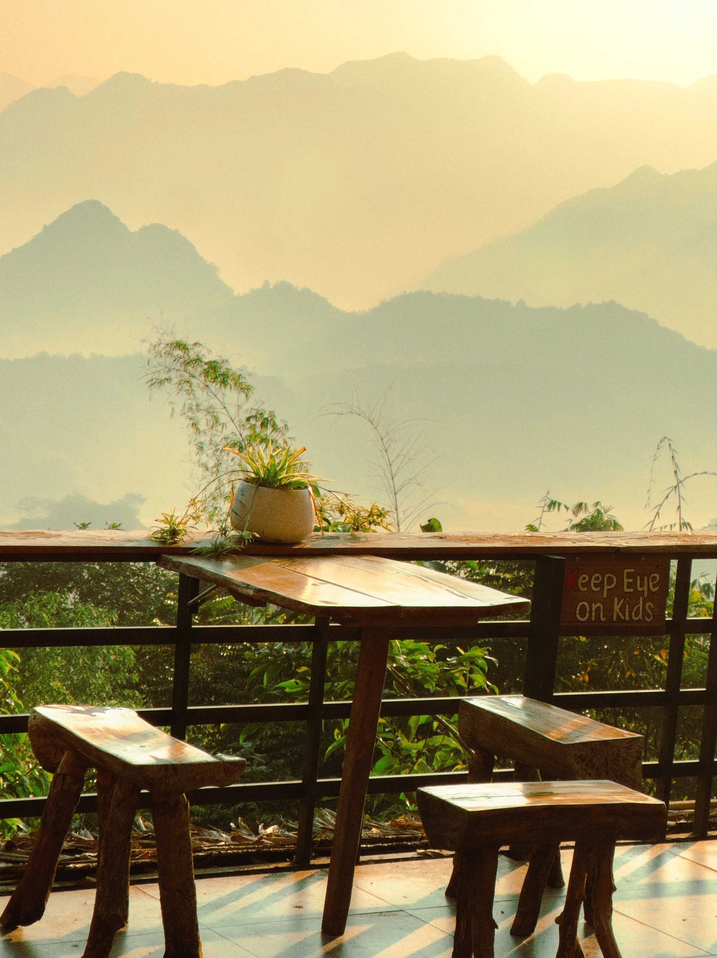 A wooden table with two wooden stools on a balcony overlooking lush greenery and mountain ranges in the distance at sunset. A potted plant sits on the table. There is a sign on the railing that says "Keep Eye on Kids."