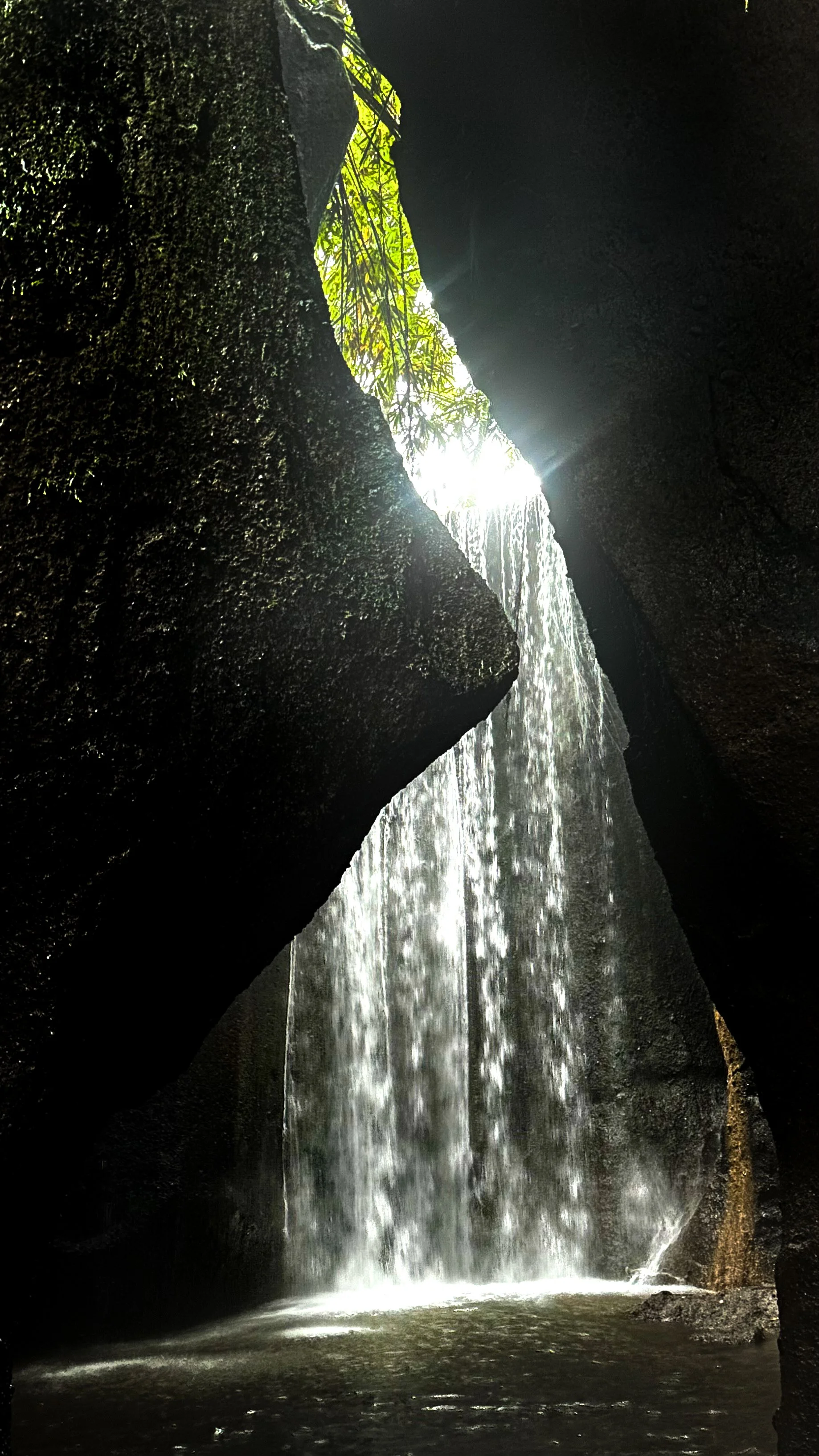 Waterfall cascading into a pool, seen from inside a narrow rocky gorge with green foliage at the top opening.