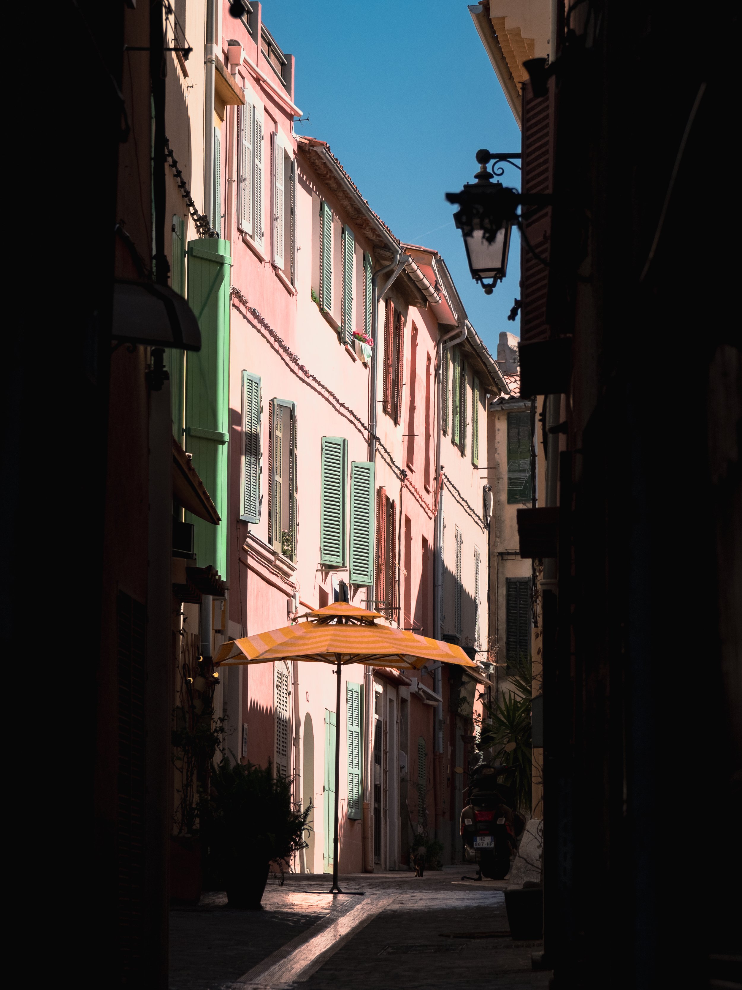 A narrow street view through a dark alleyway, with colorful pastel houses on either side, green shutters on the windows, and an orange striped umbrella on the sidewalk. A lamppost extends from the right side. The sky is blue and clear.