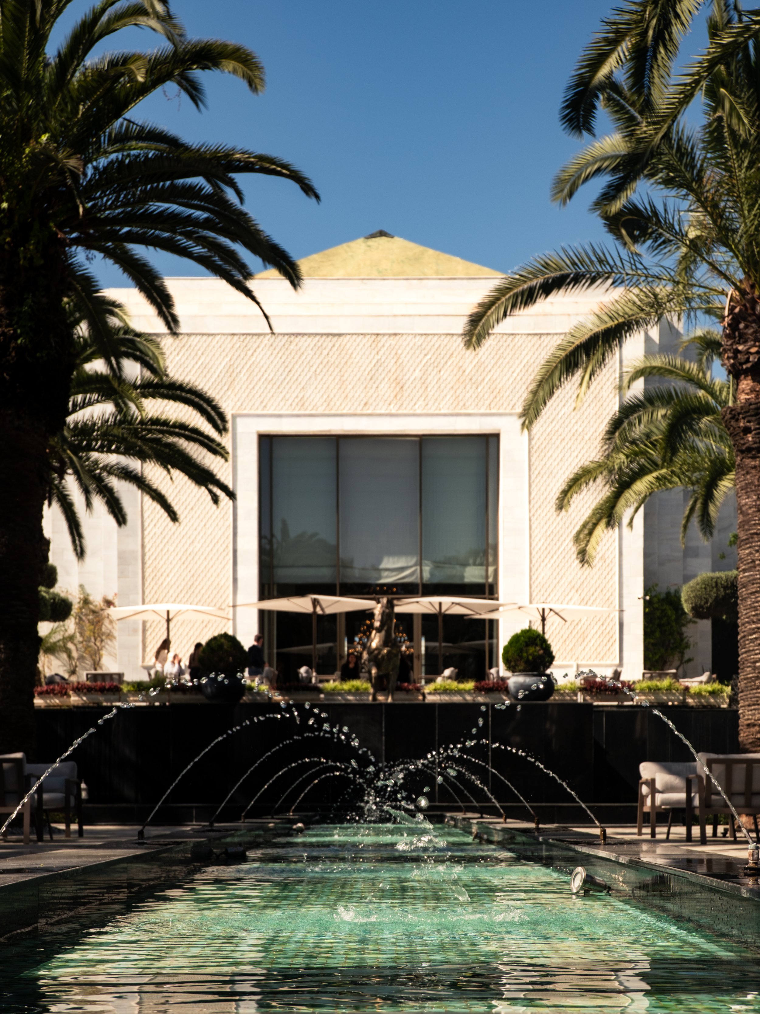 A modern building framed by palm trees, with a fountain and water feature in the foreground.