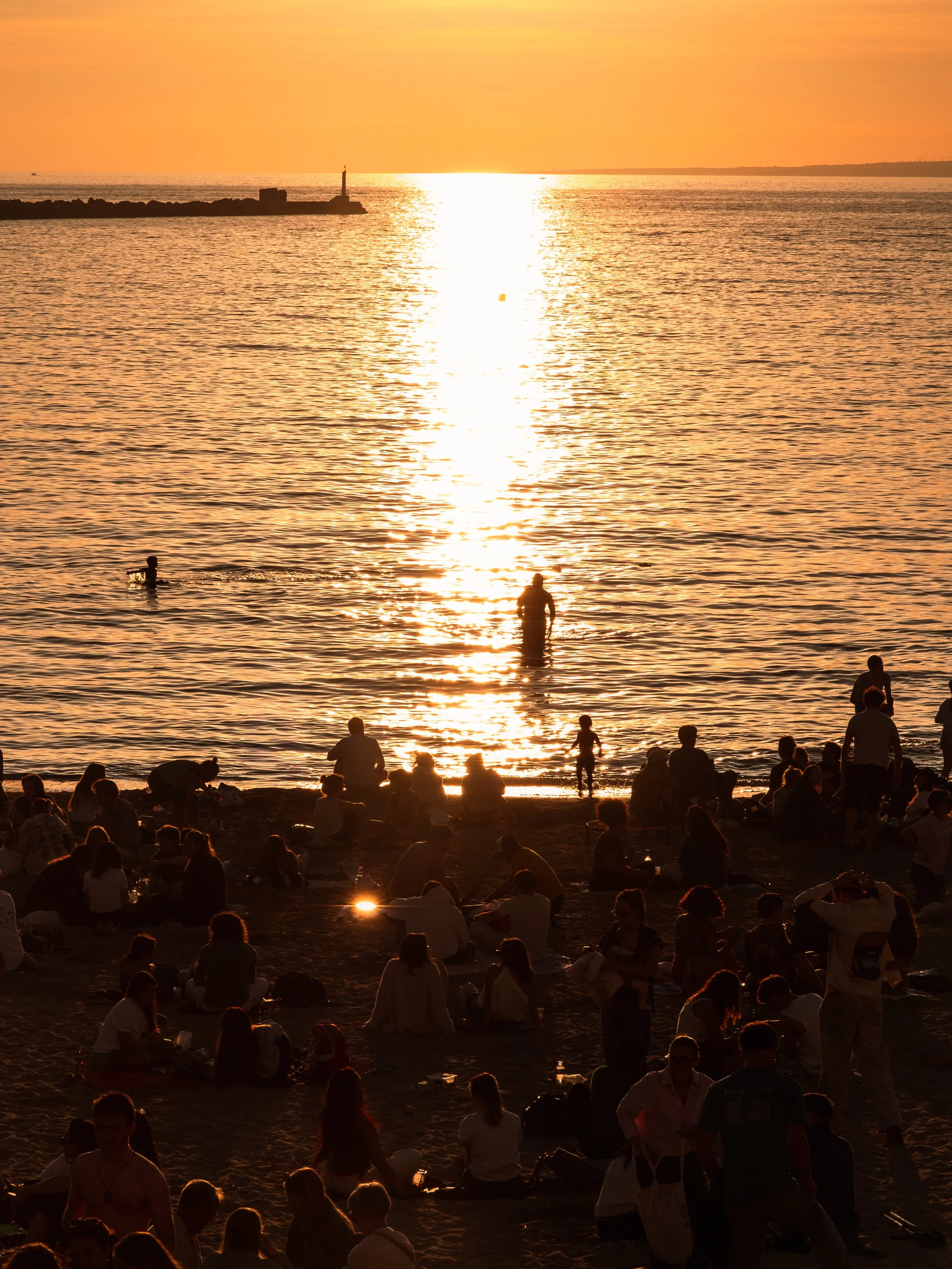 Beachgoers at sunset with silhouettes of people, water, and the sun reflecting on the ocean.