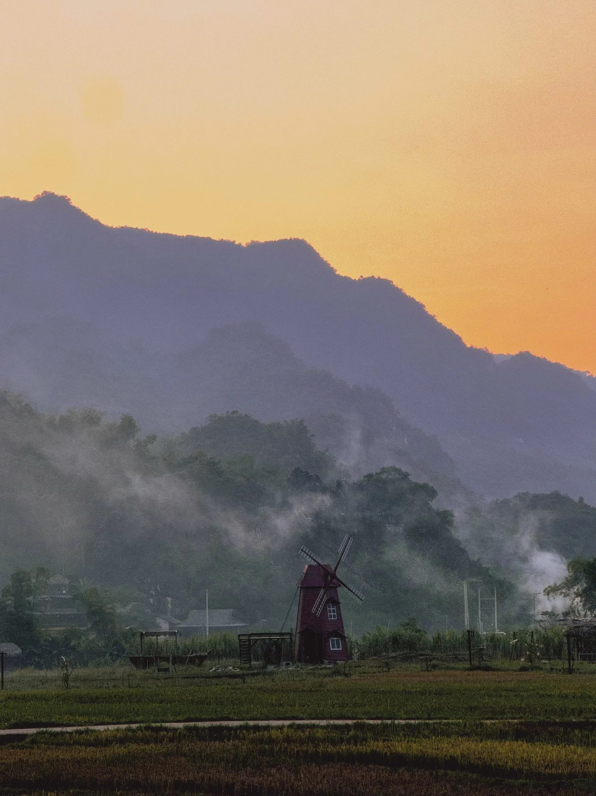 A rural landscape with a red windmill, green fields, mist in the distance, and mountains under a colorful sunset sky.