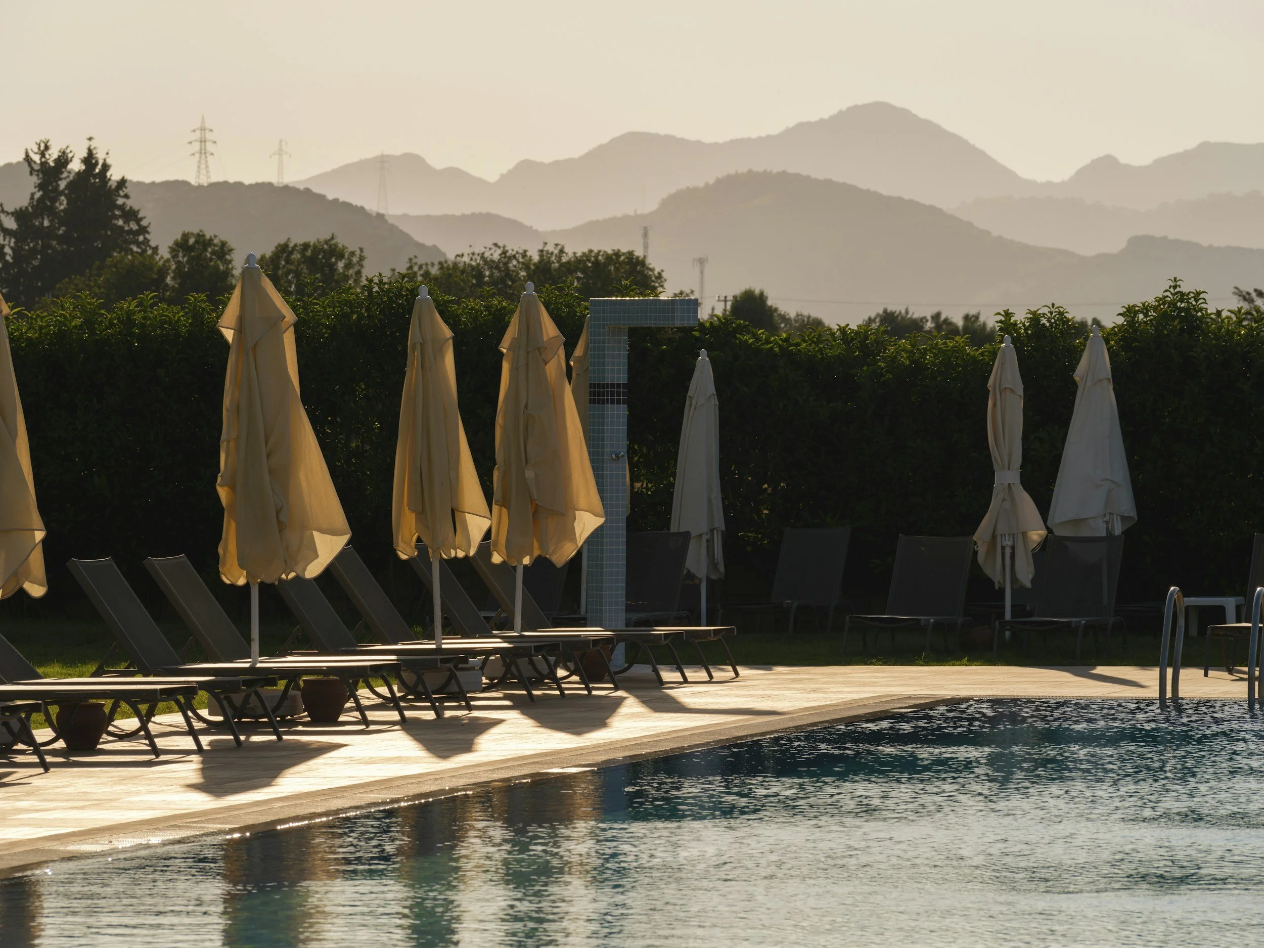 Empty poolside with closed beige umbrellas and lounge chairs, mountain landscape in the background.