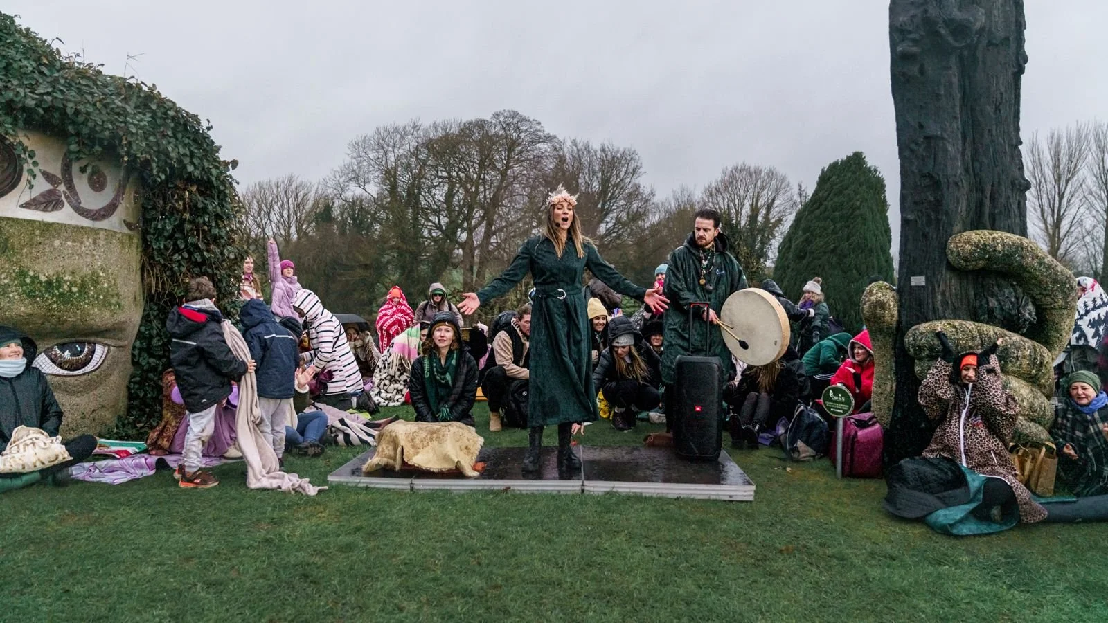 A woman in a long black coat and crown sings or speaks on a small stage outdoors, accompanied by a man with a gong. They are surrounded by a seated and standing crowd, some dressed warmly with hats and jackets, in front of an art installation with a large face sculpture and trees in the background.