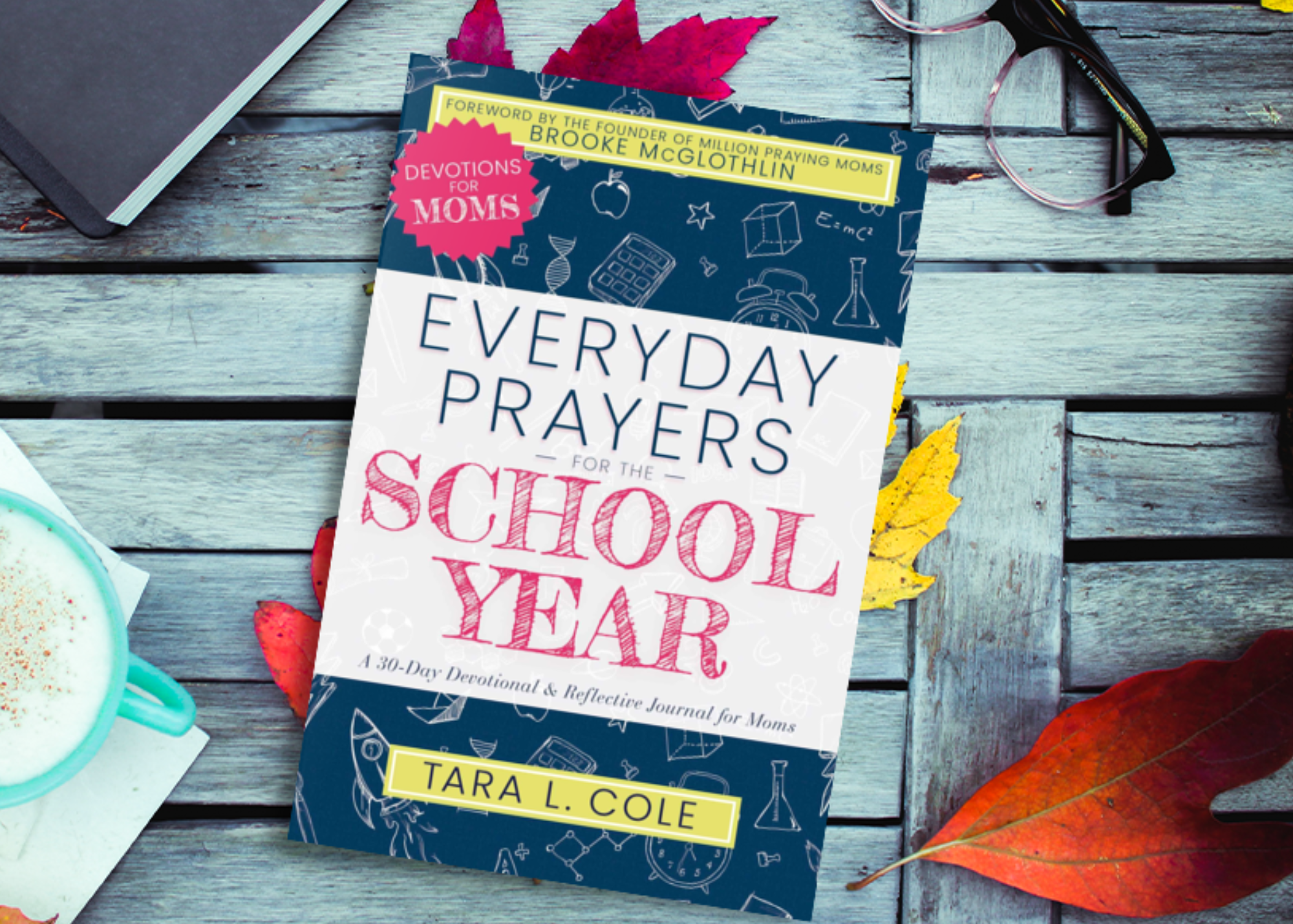 A book titled 'Everyday Prayers for the School Year' by Tara L. Cole is placed on a rustic wooden table surrounded by autumn leaves, a pair of glasses, a notebook, and a mug of frothy beverage.
