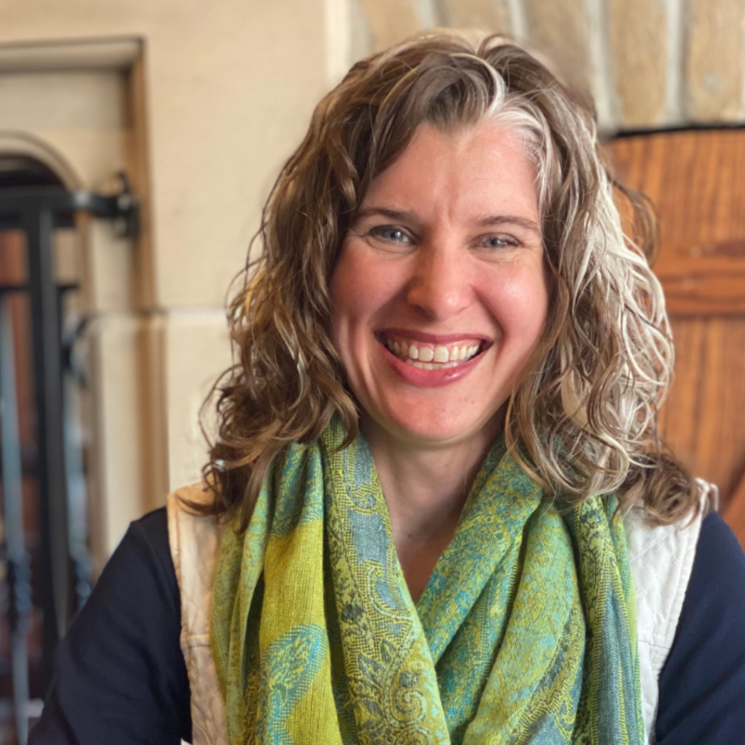 Close-up of a Tara L. Cole with curly, shoulder-length hair wearing a green patterned scarf and a white vest, in a cozy indoor setting.