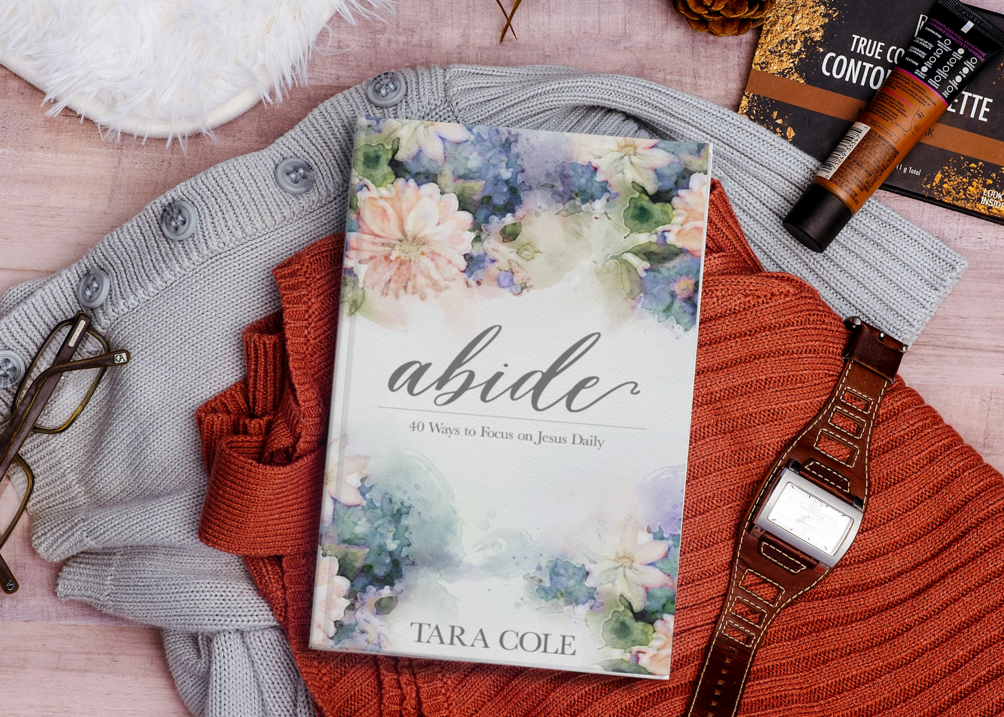 A book titled 'Abide: 40 Ways to Focus on Jesus Daily' by Tara Cole, placed on colorful clothing. Surrounding items include a pair of eyeglasses, a wristwatch, a smartphone, a tube of lotion or cosmetic product, a gray knit sweater, an orange knit ga