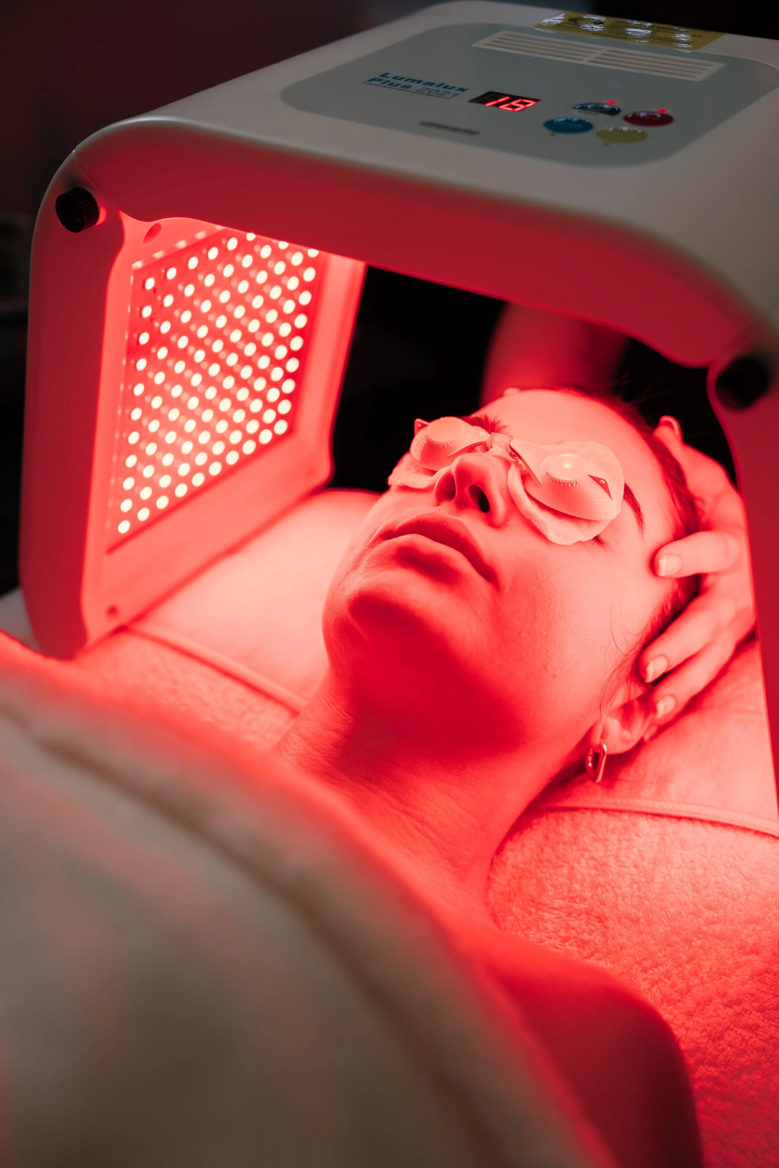 A woman lying on a treatment bed undergoing red light therapy, wearing eye protection, with a red light device positioned above her face.