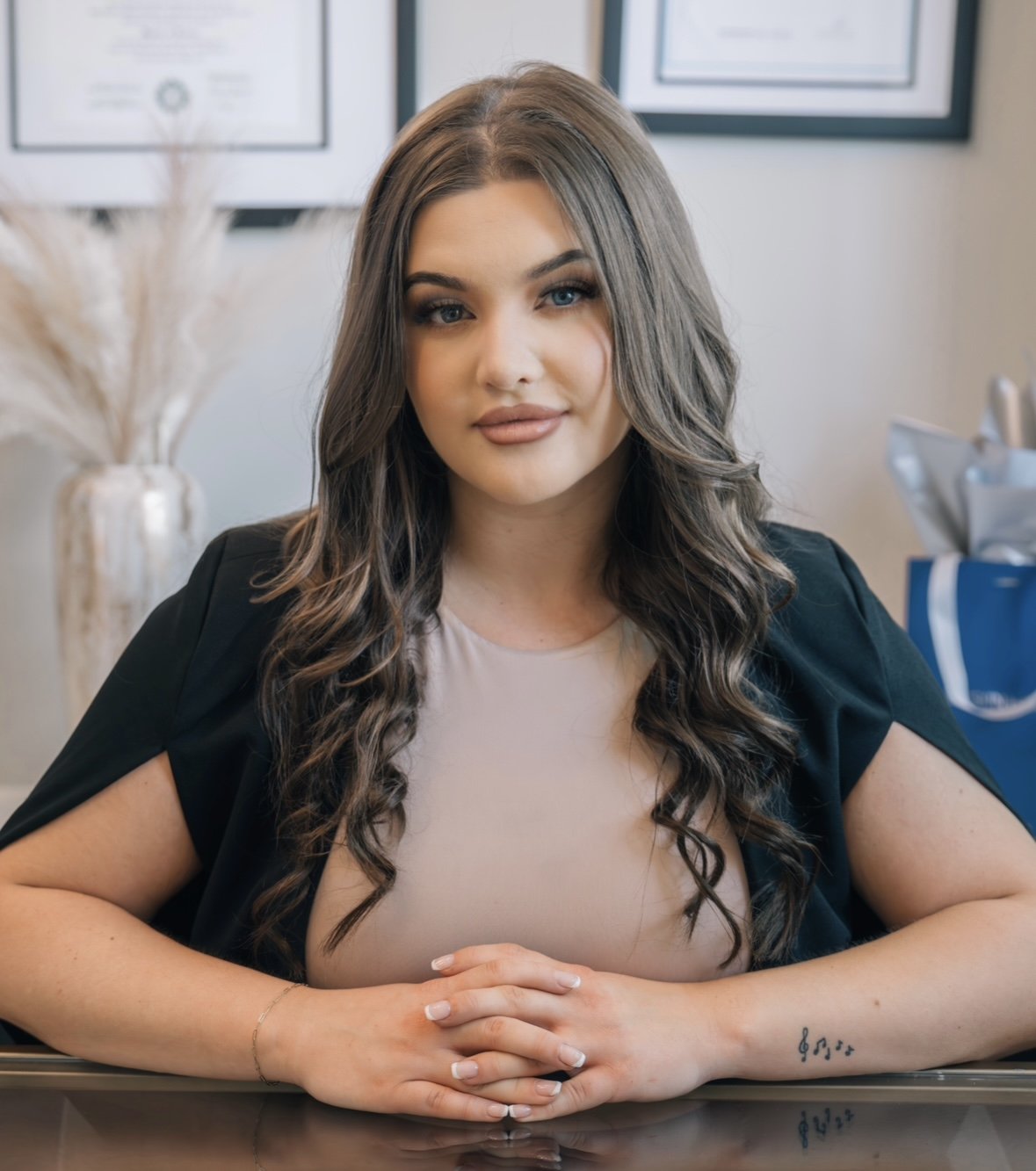 A young woman with long wavy brown hair and blue eyes sitting at a desk, smiling slightly. She is wearing a beige top and a black blazer, with a small tattoo on her right wrist. The background shows framed certificates on the wall and decorative dried plants in a vase.