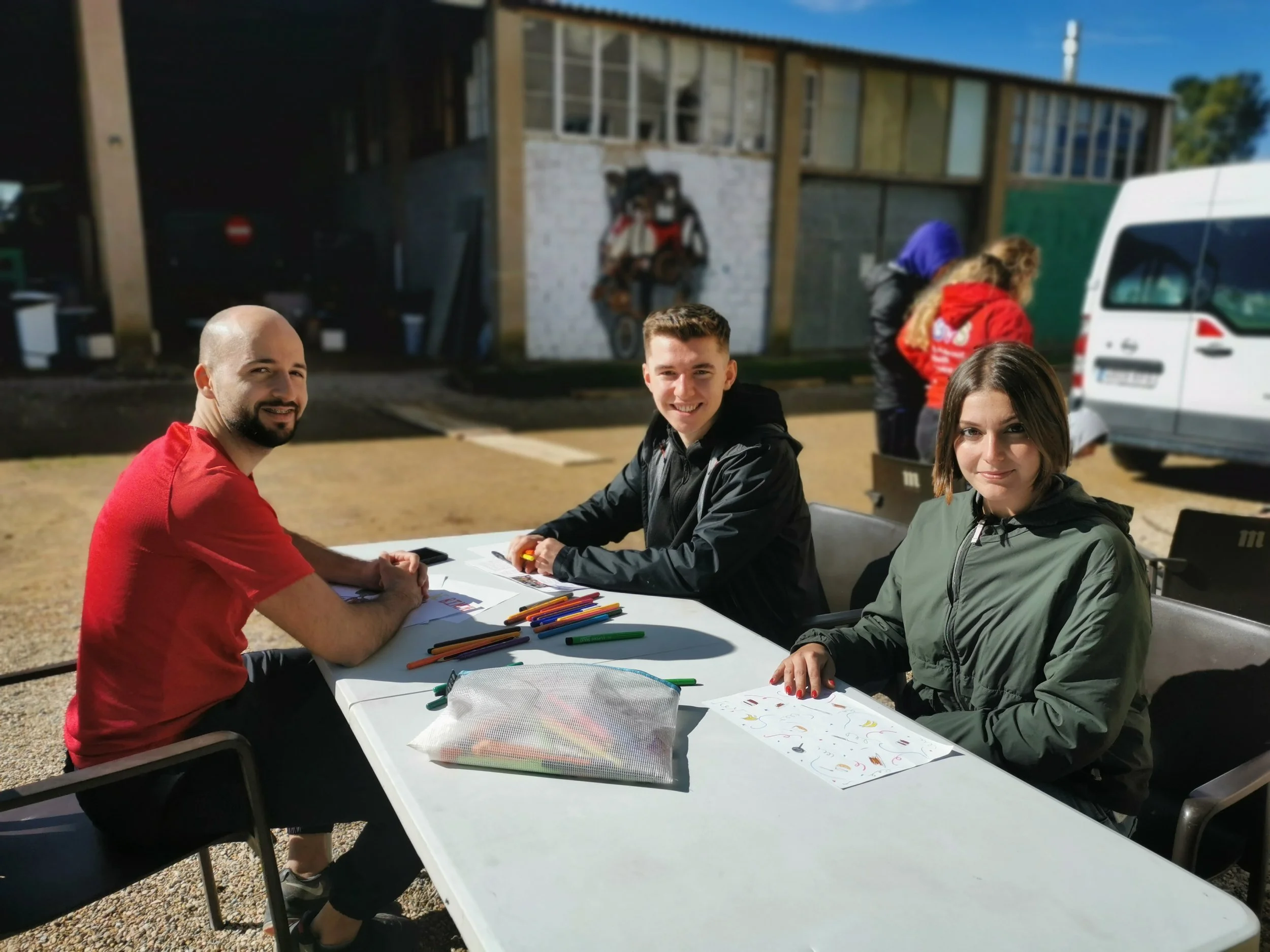 Tres jóvenes trabajando entorno a una mesa miran a cámara y sonríen. 