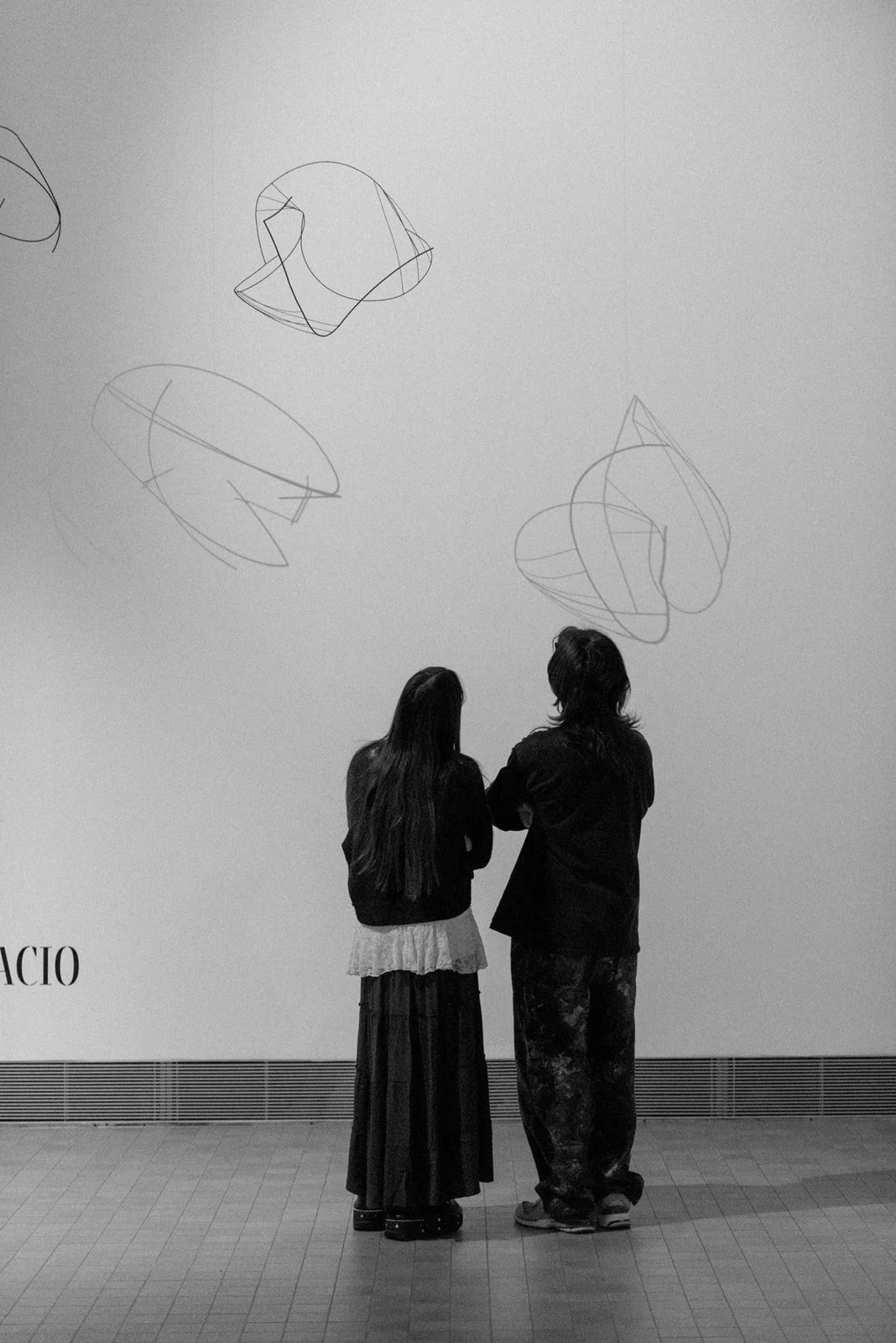 Imagen en blanco y negro. Una chica y un chico de espaldas miran a esculturas que penden del techo y que se elevan por encima de sus cabezas, proyectando sobras sobre la pared.