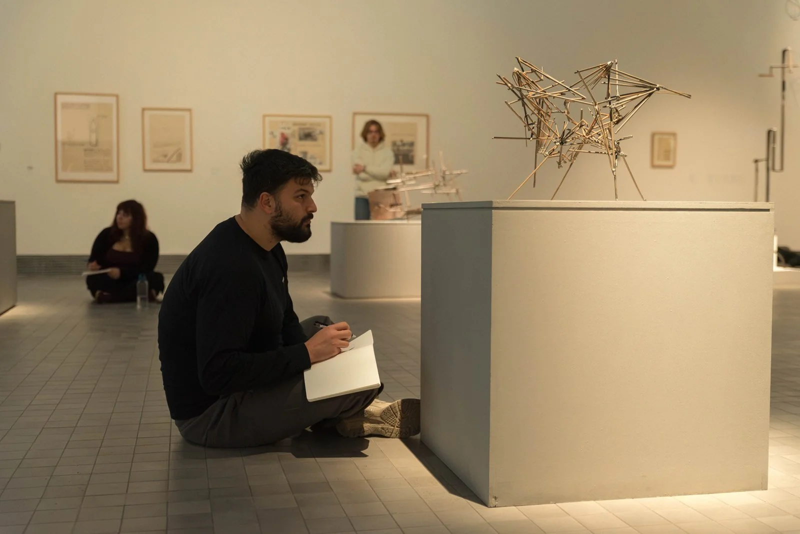 Un hombre está sentado en suelo de un museo, mientras contempla una escultura que está sobre una peana. Tiene un cuaderno en sus manos y sostiene un bolígrafo.
