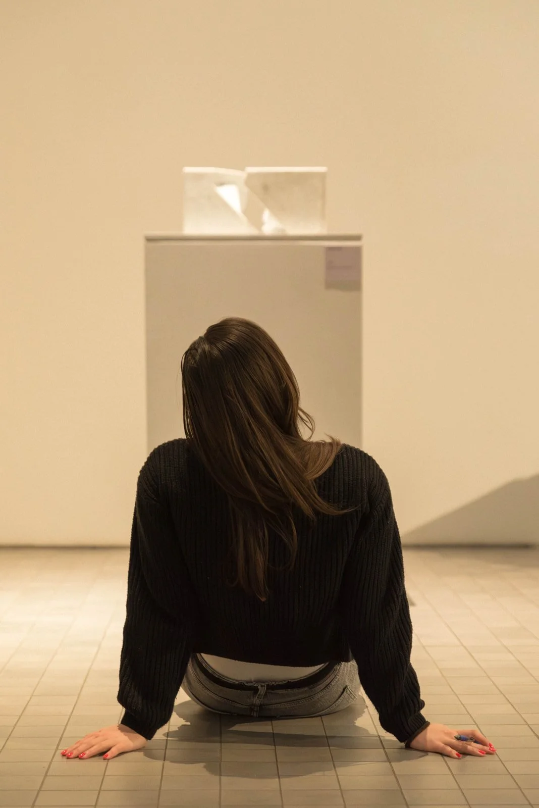 Chica de espaldas, sentada en el suelo y contemplando una obra de arte que está sobre una peana y que se ve justo detrás de ella, por encima de su cabeza.